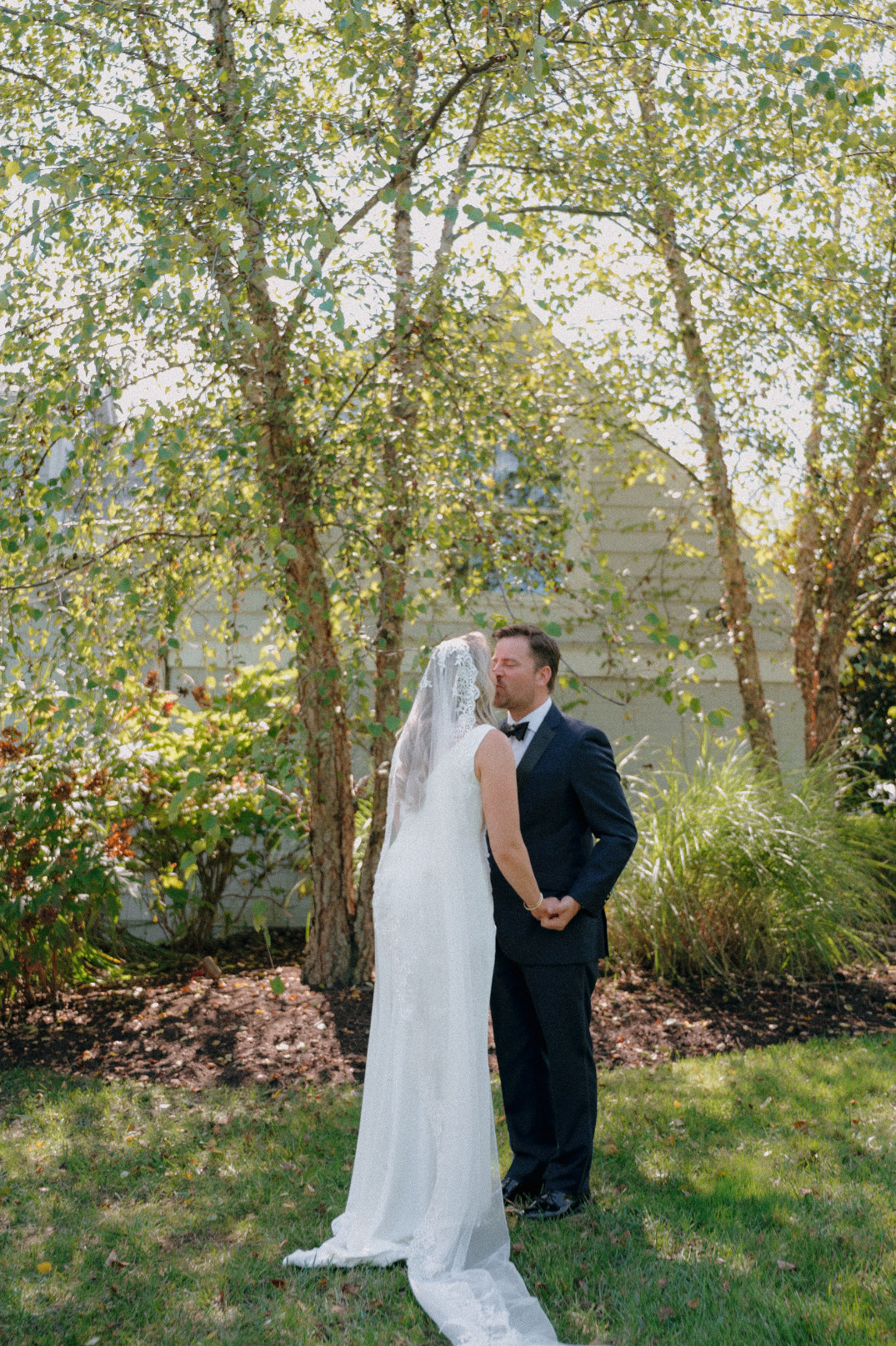 A bride and groom share a kiss in a sunlit garden. She wears a long, white dress and veil; he wears a dark suit. They stand surrounded by trees in Ohio