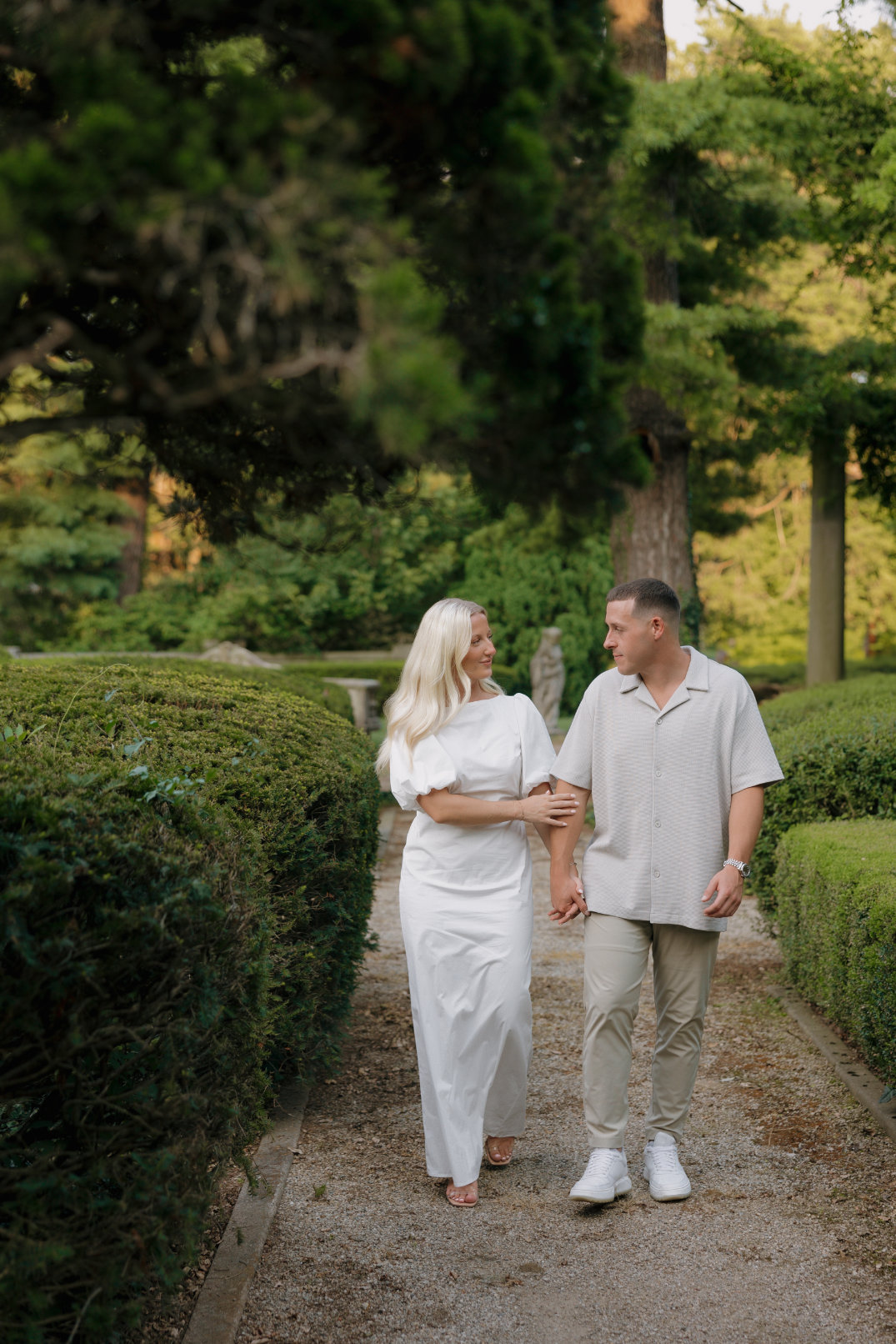 The couple walking hand in hand along a garden path surrounded by greenery.