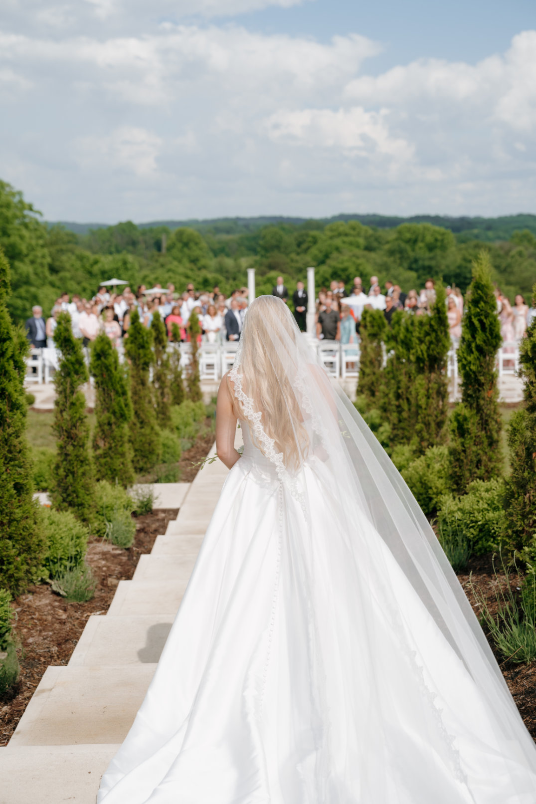 Bride in a flowing white gown walking down an outdoor aisle lined with greenery toward guests. The sky is blue and the scene is serene and joyful.