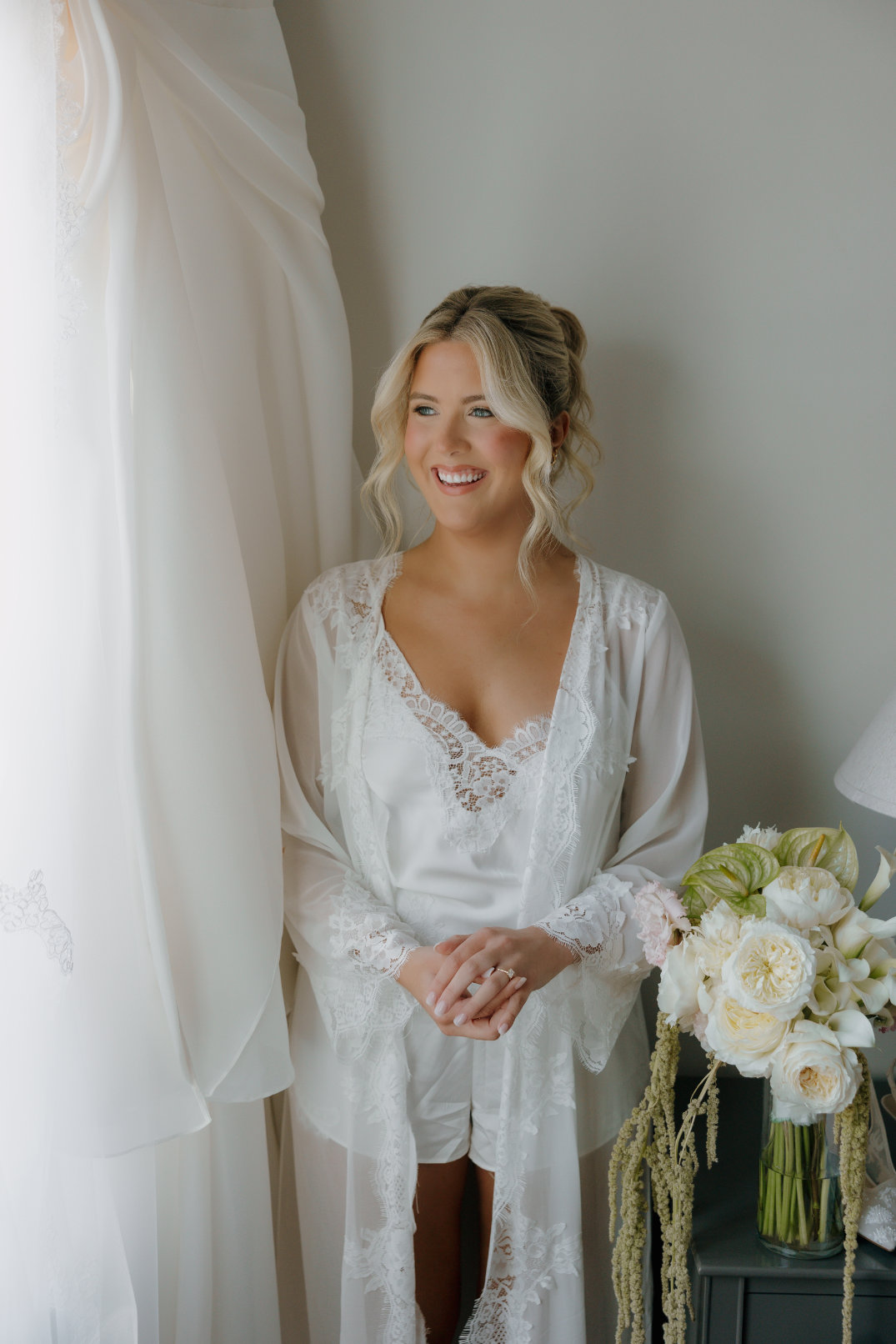 A smiling woman in a white lace robe stands by a window with soft curtains. A bouquet of white and pale yellow roses adds elegance and warmth to the scene at The Orchid House Winery.