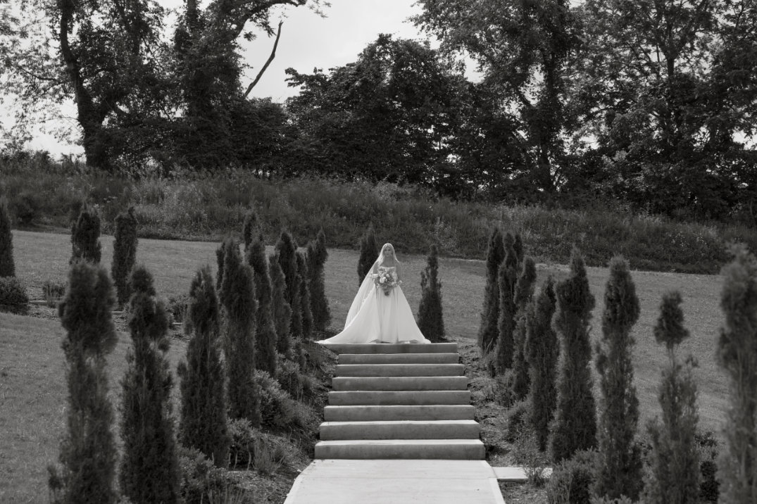 A bride in a flowing gown descends stone steps bordered by tall, narrow trees. The scene is peaceful, set amidst lush greenery under a soft, overcast sky.