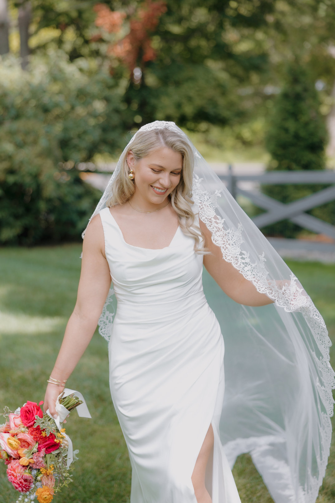 A smiling bride in a white dress with a lace veil holds a vibrant flower bouquet. She stands on green grass with a wooden fence and trees in the background.