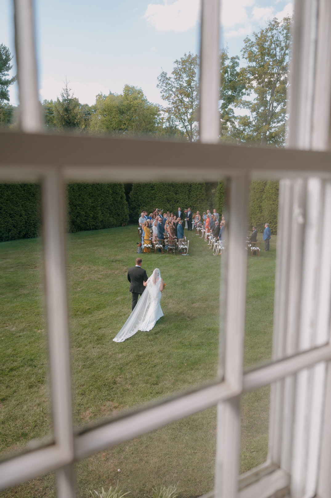 View through a window of a bride and groom walking on a lawn towards a seated wedding crowd surrounded by trees, under a bright blue sky.
