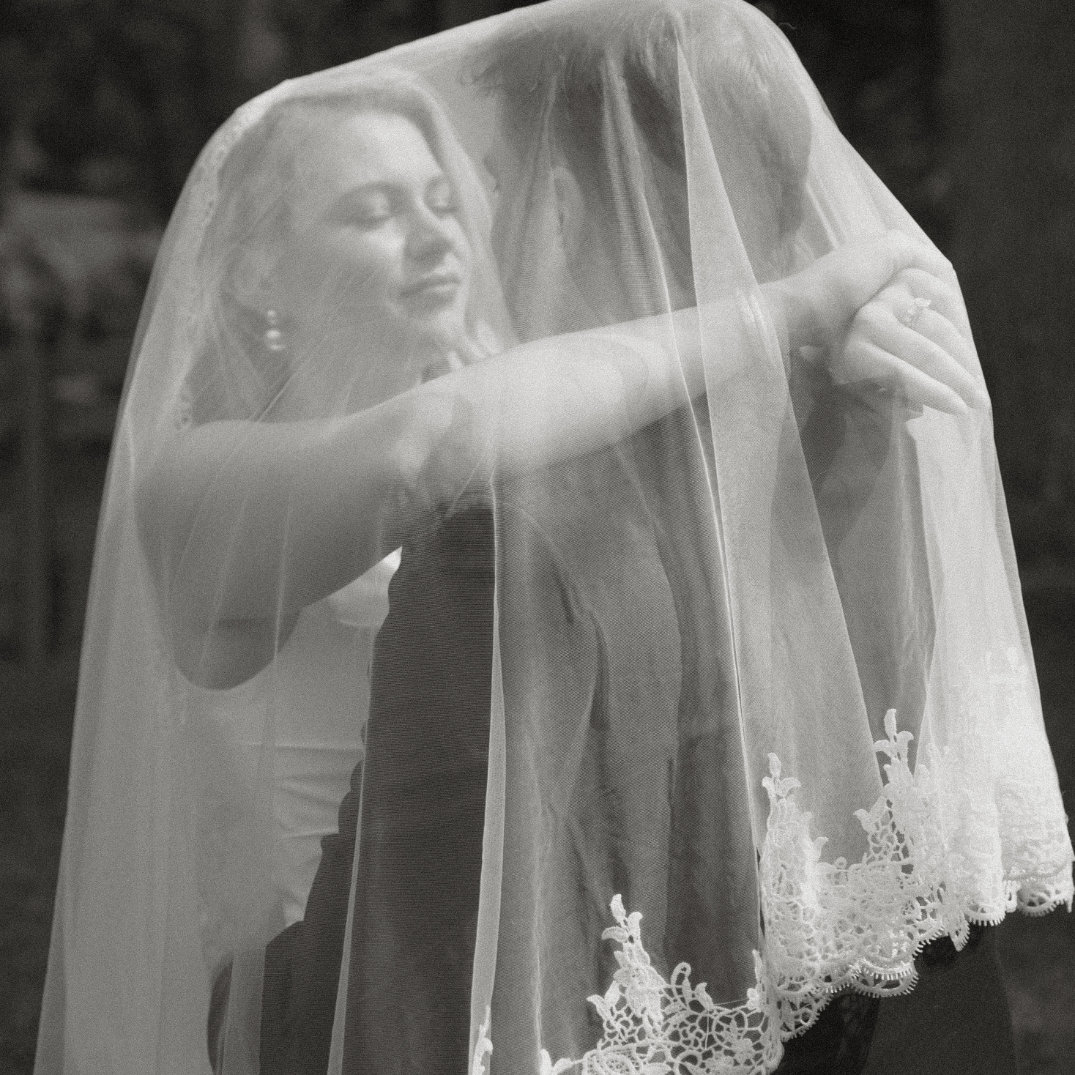A bride and groom share an intimate embrace under a lace-edged veil, captured in black and white. Their expressions convey joy and tenderness.