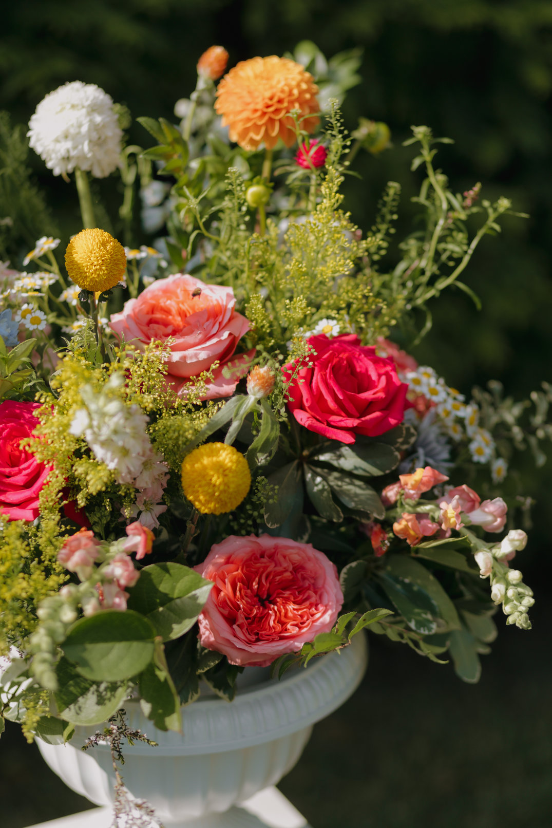A vibrant floral arrangement in a white vase with red, pink, and orange roses, yellow and white blooms, greenery, and a lush, garden backdrop.