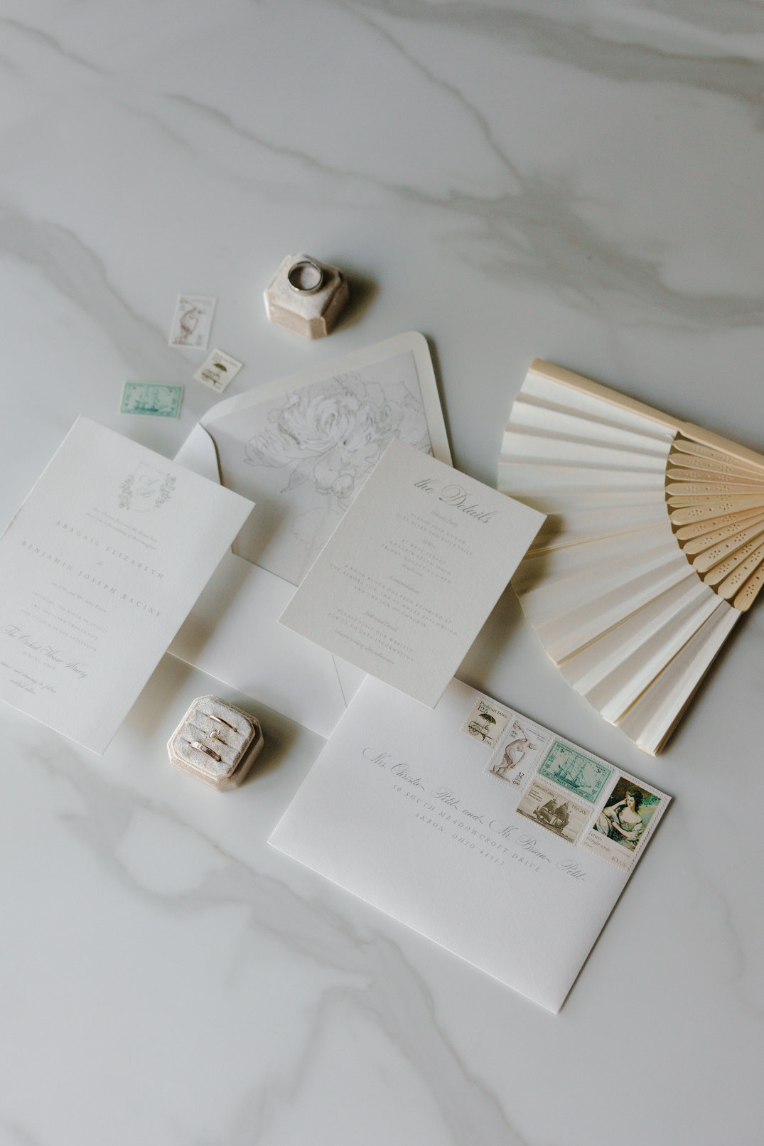 Elegant flat lay of wedding stationery on a marble surface, featuring invitations, envelopes with vintage stamps, a wooden fan, ink stamps, and a ring box.
