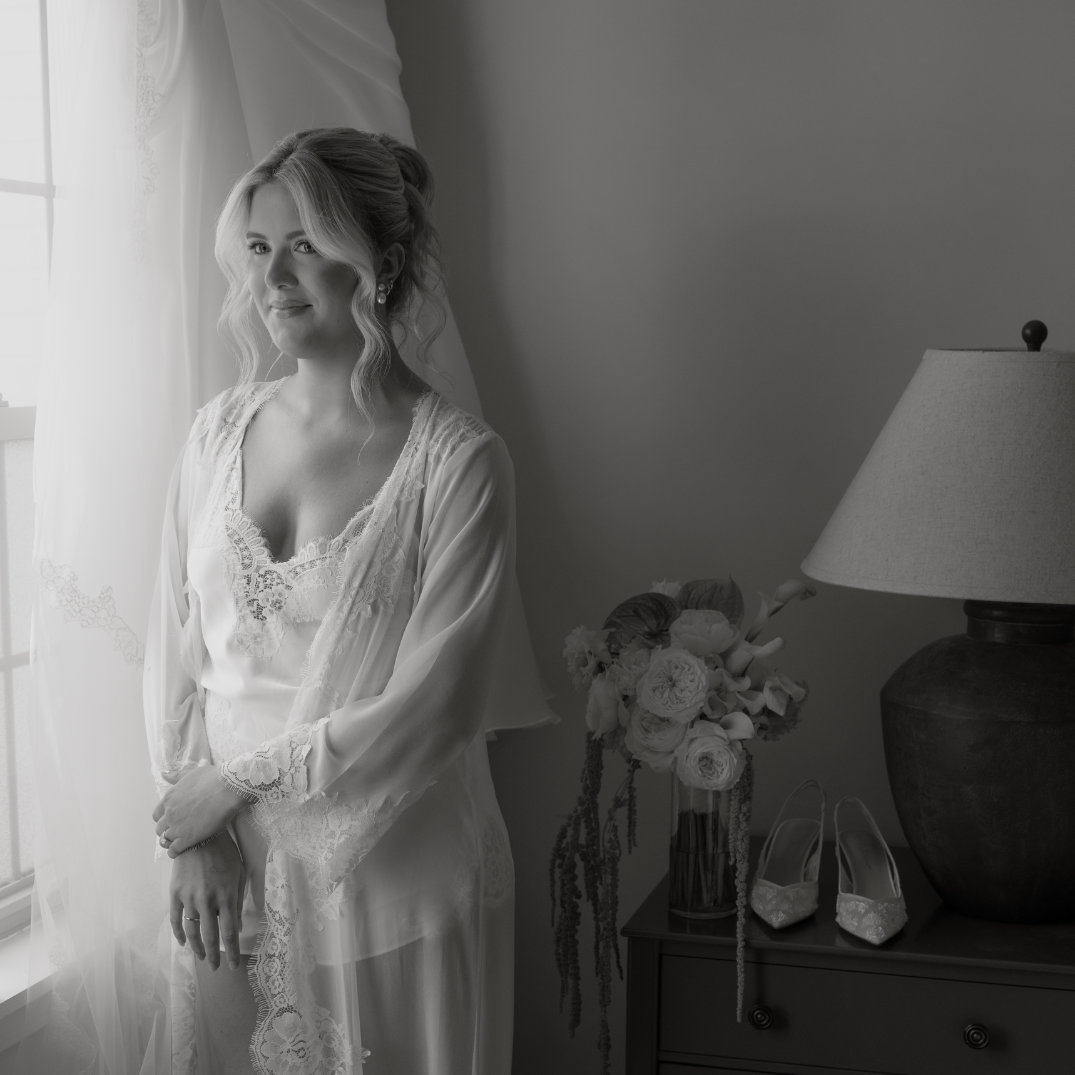 Ohio Bride in lacy robe gazes thoughtfully out the window, standing beside a lamp and flowers on a dresser. Her shoes are placed neatly below.