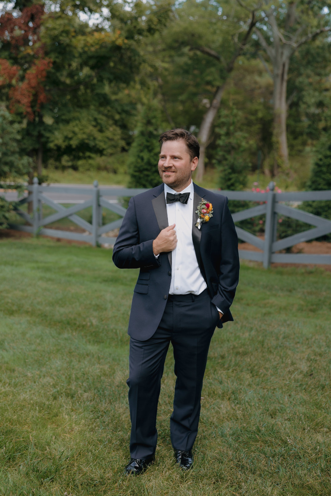 A man in a dark suit with a bow tie stands confidently on grass, one hand in his pocket. He's smiling, with trees and a wooden fence behind him.