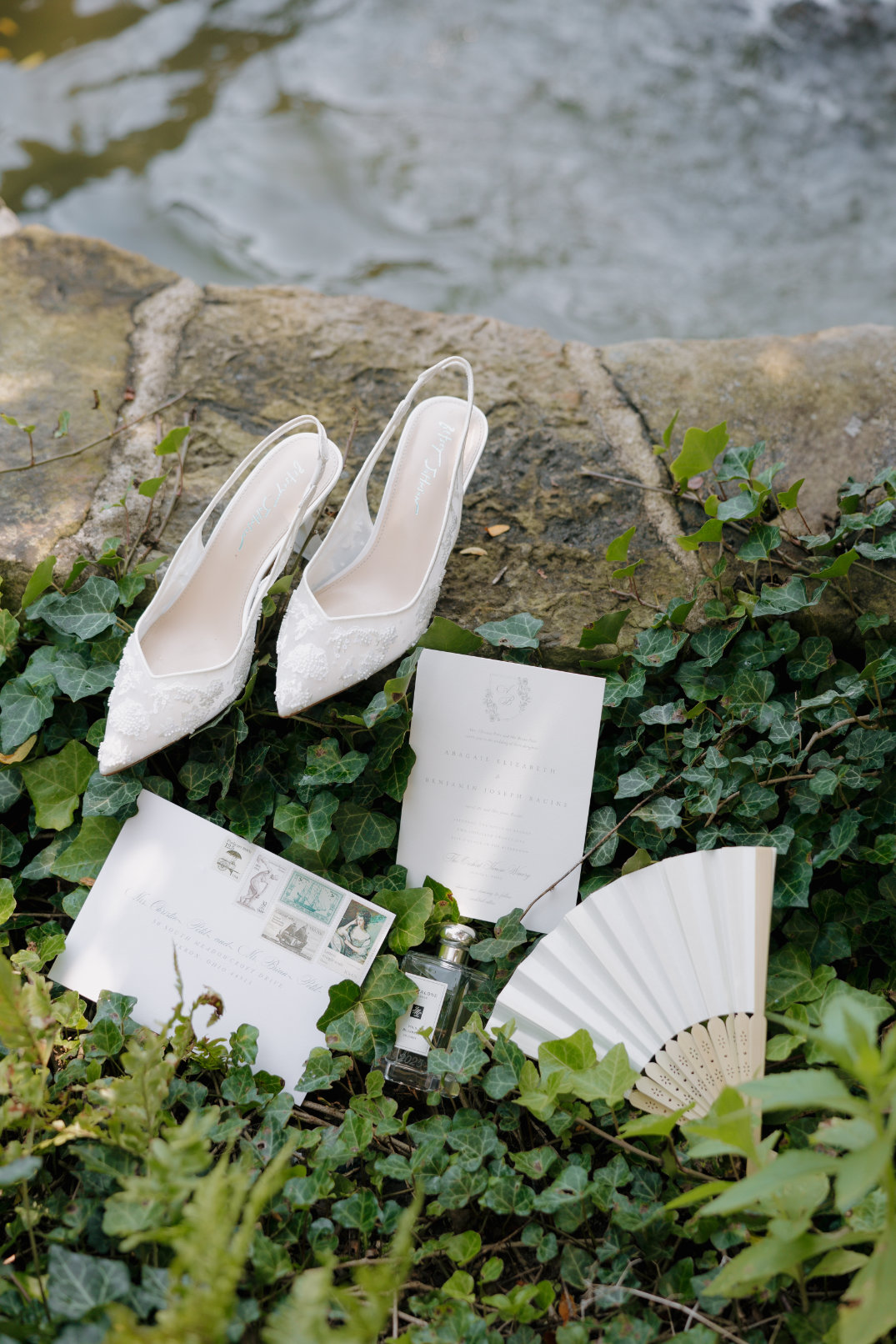 Elegant white bridal shoes, a fan, wedding invitations, and a perfume bottle are arranged on green ivy by a stone path, evoking a romantic setting.