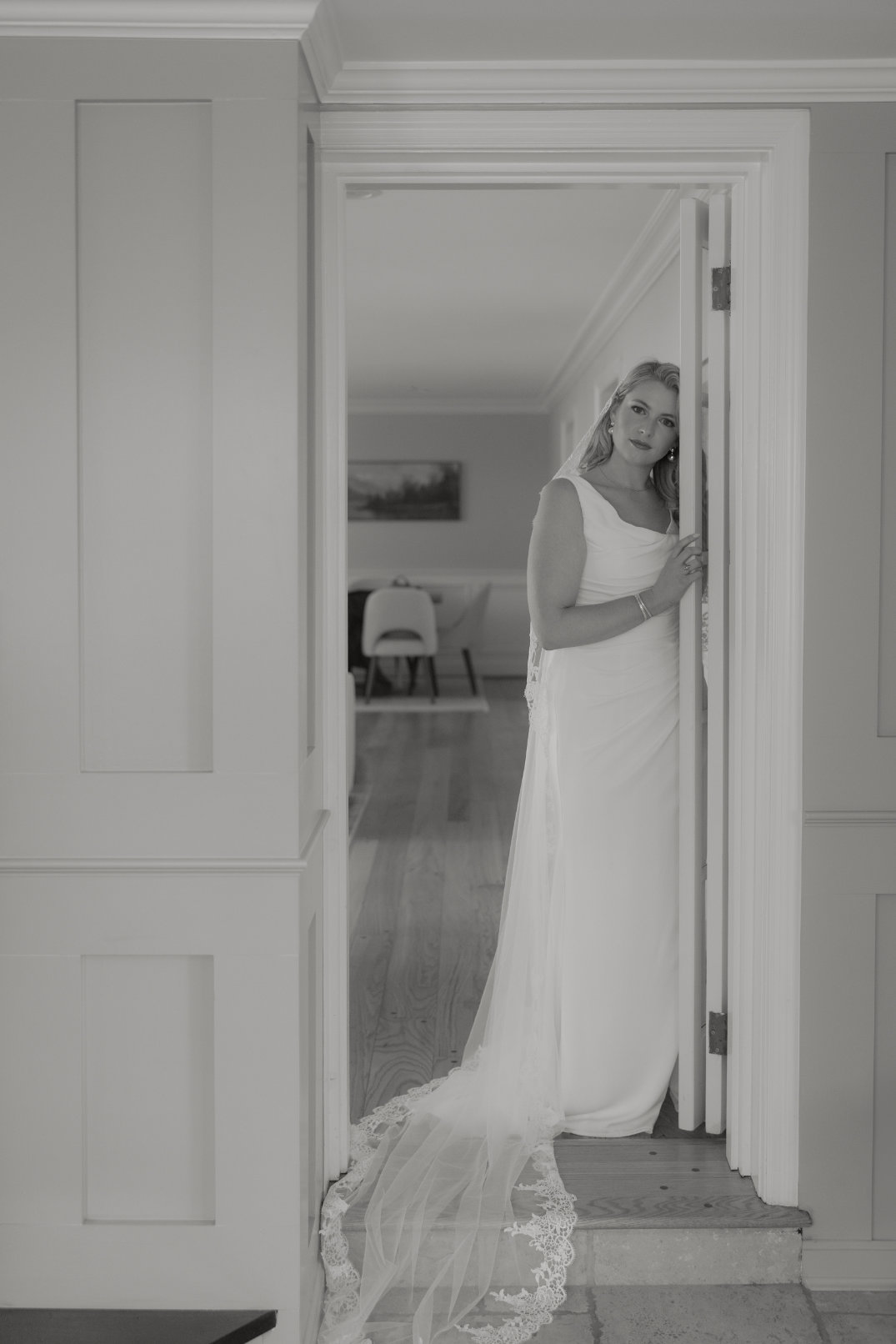 Bride in an elegant gown stands in a doorway, gazing gently with a serene expression. Her long veil trails behind, and a dimly lit room is visible in the background.