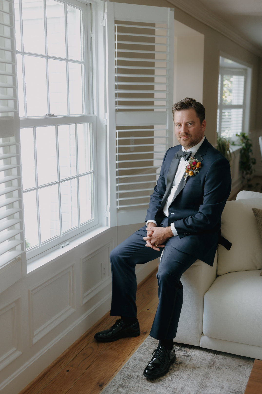 A man in a navy suit with a floral boutonniere sits on a sofa by a window with white blinds. The setting is bright and elegant, conveying a calm and formal tone.