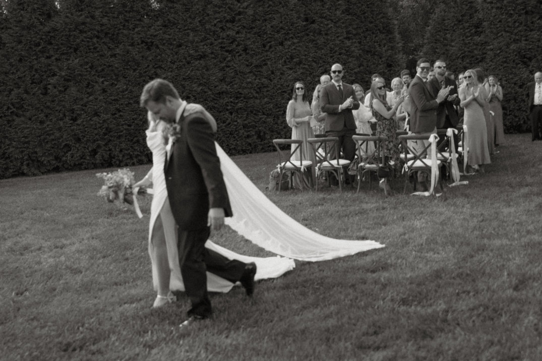 A bride and groom walk on grass, the bride holding a bouquet and wearing a long veil. Guests stand and clap in a joyful outdoor setting. Black and white image.