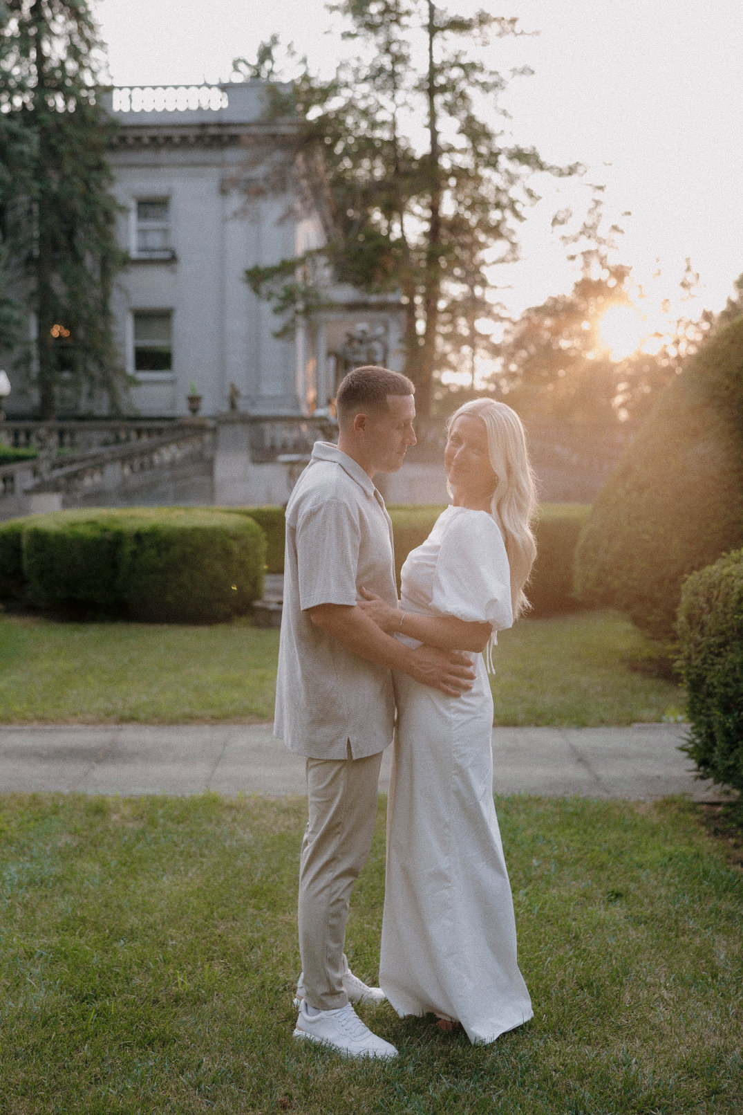 The couple embracing in warm sunset light with trees and stone architecture behind them.
