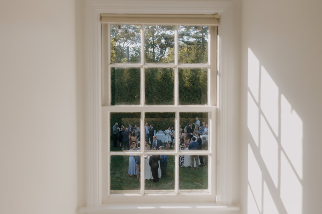 View through a window showing a garden party. Groups of people in formal wear socialize on a lawn, surrounded by greenery, under a bright sky.