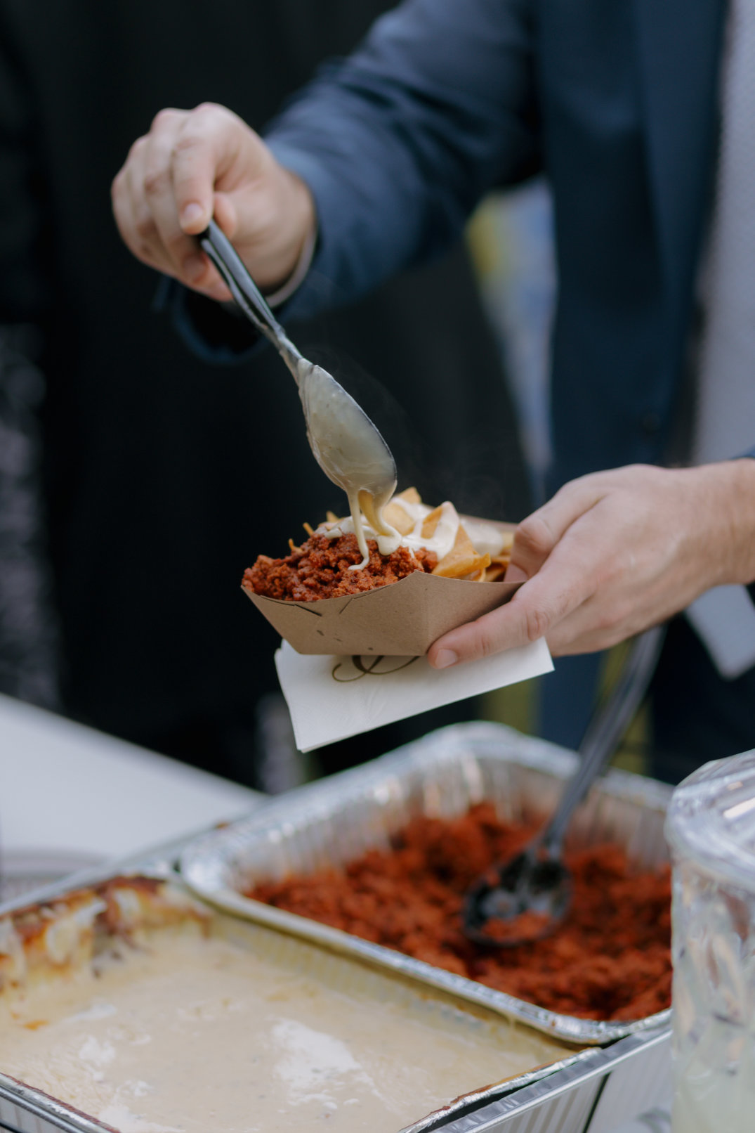 A person holds a paper tray with tortilla chips and crumbled meat, drizzling creamy sauce from a spoon. Metal trays with food are visible in the background.