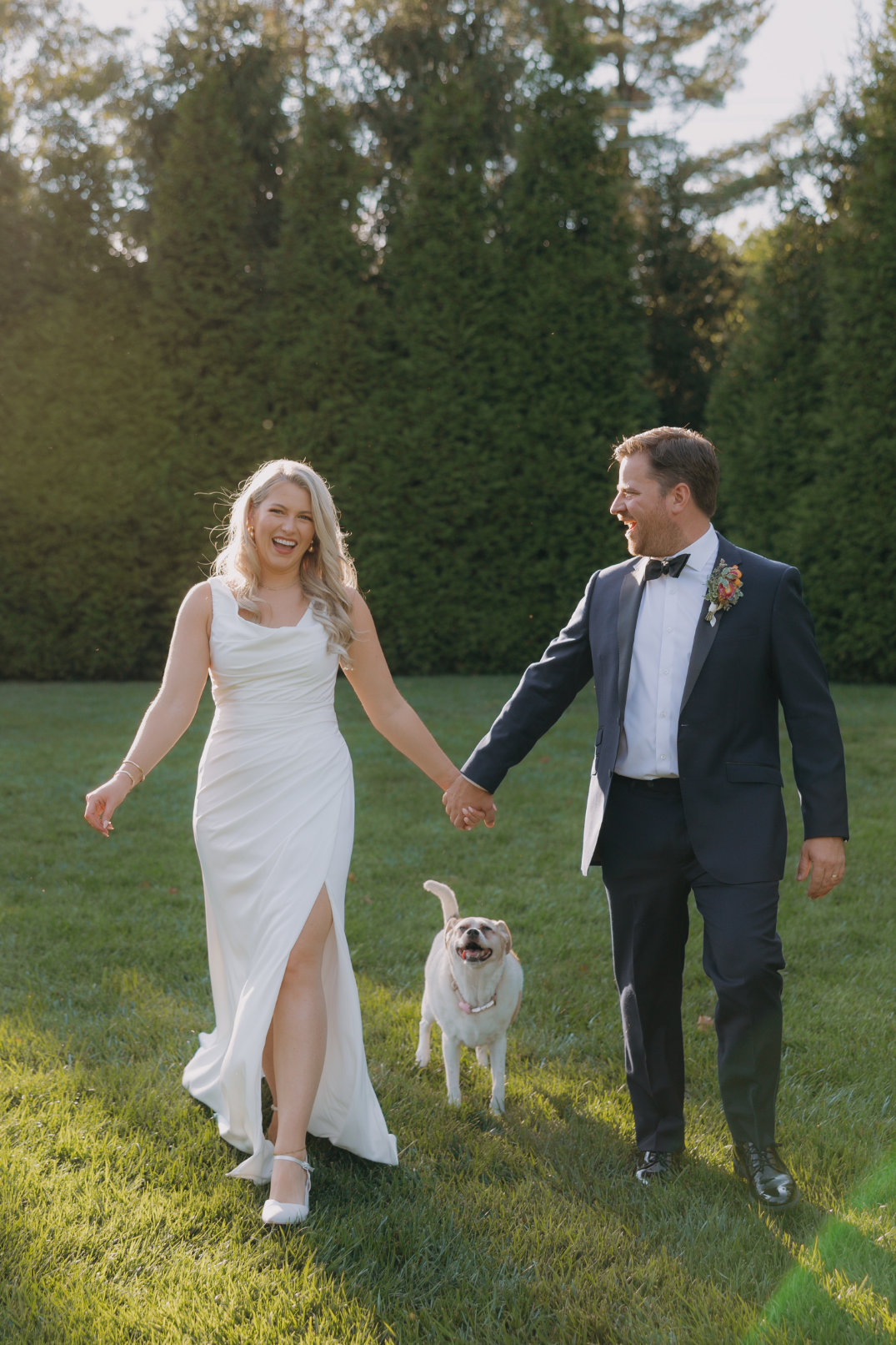 A happy couple in wedding attire walks hand in hand on grass, accompanied by a small, joyful dog. Sunlight filters through trees, creating a warm, cheerful atmosphere.