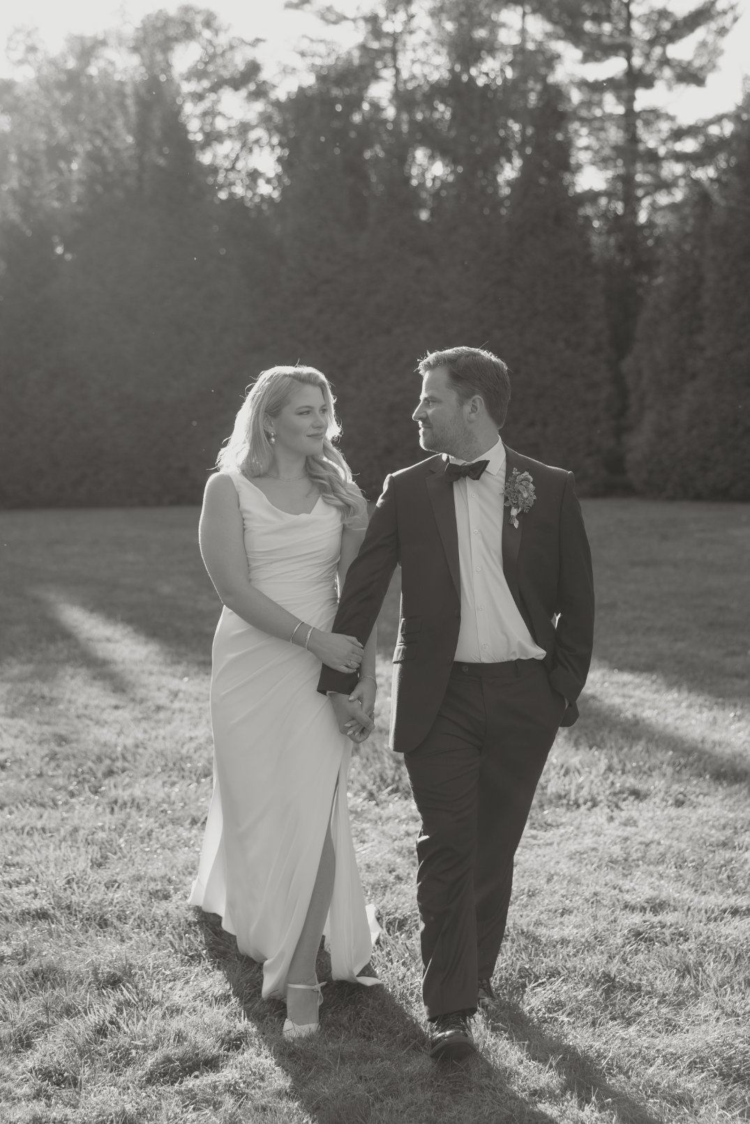 A Cincinnati couple walks hand in hand on a sunlit lawn. She wears a flowing white dress, and he is in a suit with a bow tie, sharing a joyful moment.