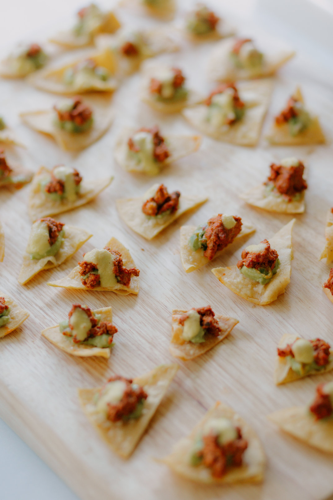 Triangular chips topped with guacamole and seasoned meat are neatly arranged on a wooden board. The image conveys a tasty and inviting snack display.