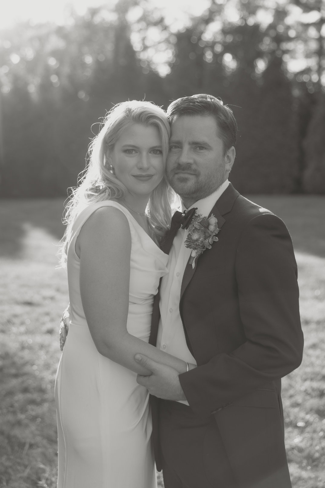 A newlywed couple embraces in a garden, dressed in wedding attire. Sunlight filters through trees, creating a warm, romantic ambiance. Black and white tones.