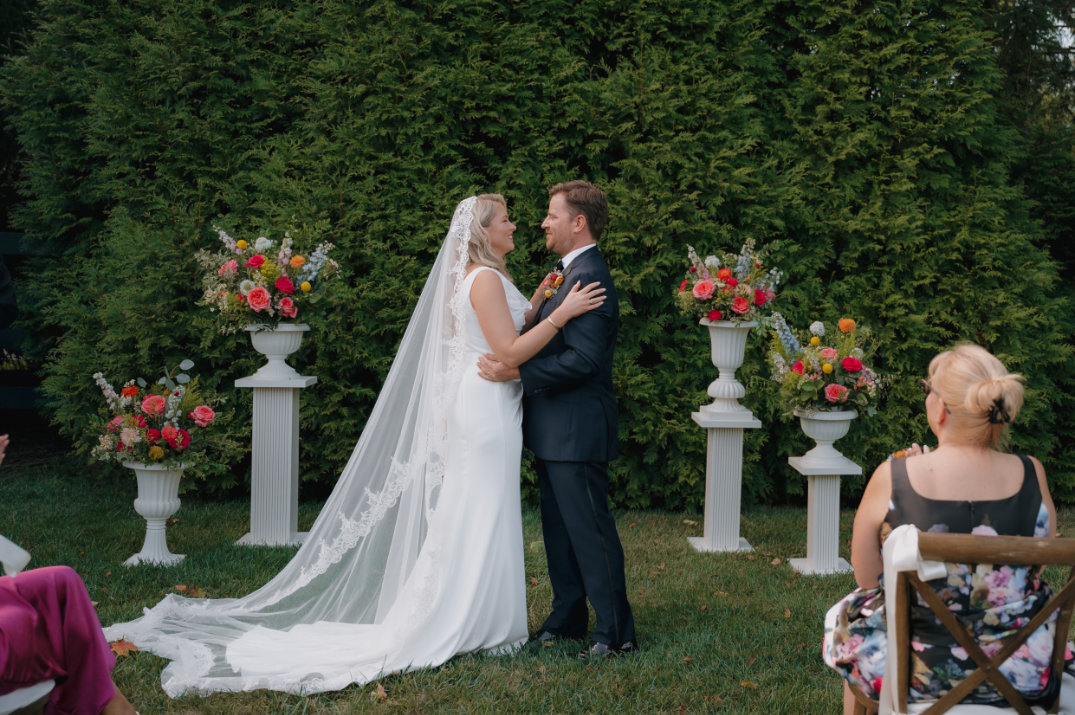 A bride and groom embrace outdoors before lush greenery. Floral arrangements in white vases surround them. Guests in colorful attire watch.