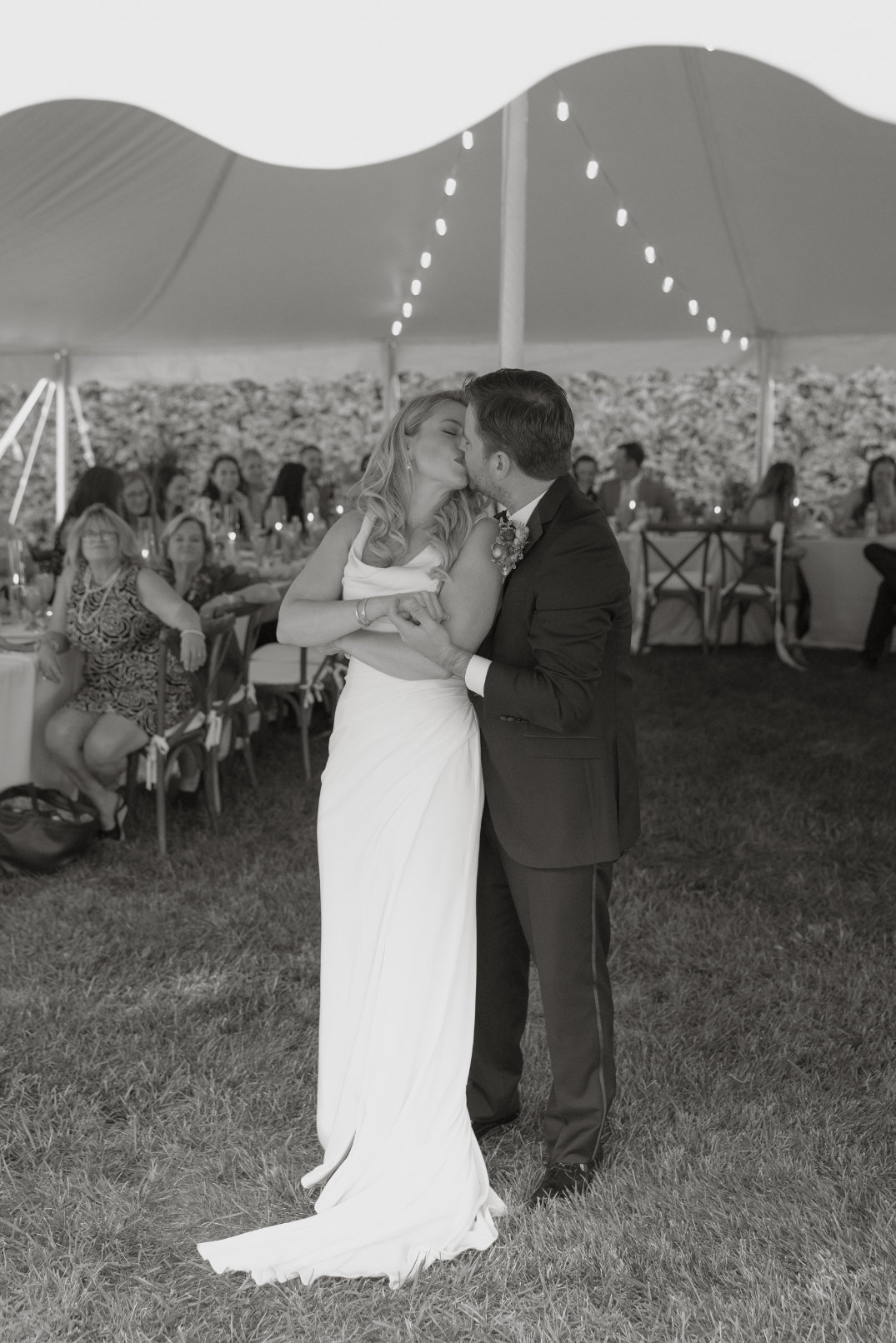 Bride and groom share a loving kiss under a tent adorned with string lights during their wedding reception, surrounded by smiling guests. Black and white photo.