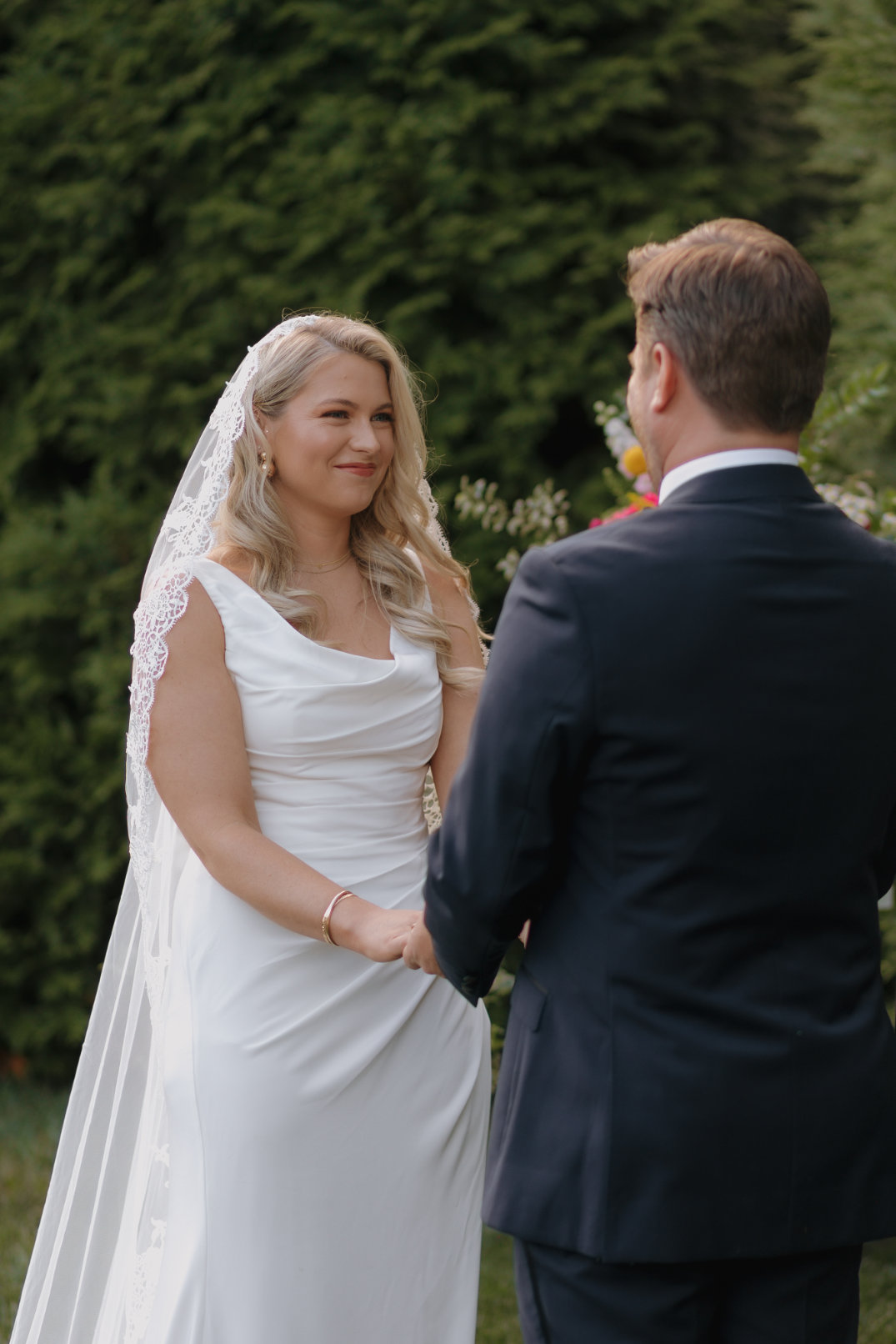 A bride and groom stand outdoors, holding hands facing each other. The bride wears a white dress and lace veil, smiling softly. Lush greenery surrounds them.