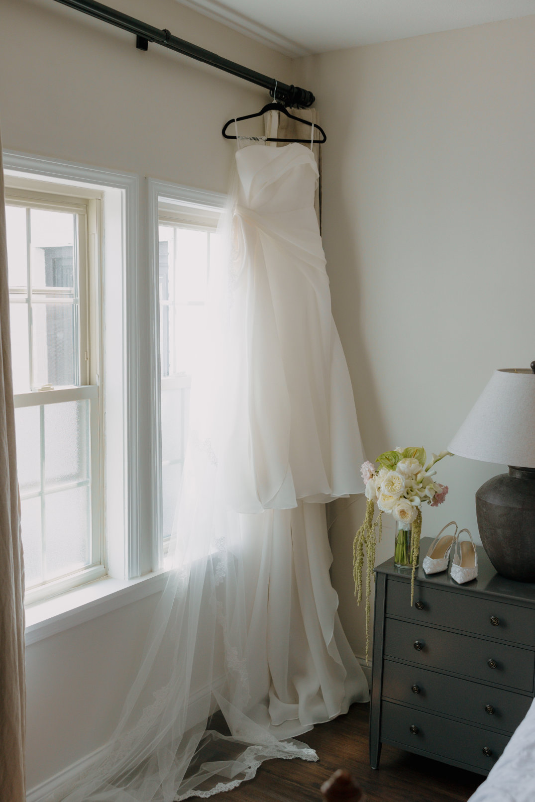 A flowing white wedding dress for a Cleveland bride, hangs near a sunny window. Beside it, a nightstand holds a vase of flowers, a pair of heels, and a lamp, creating an elegant, serene atmosphere.