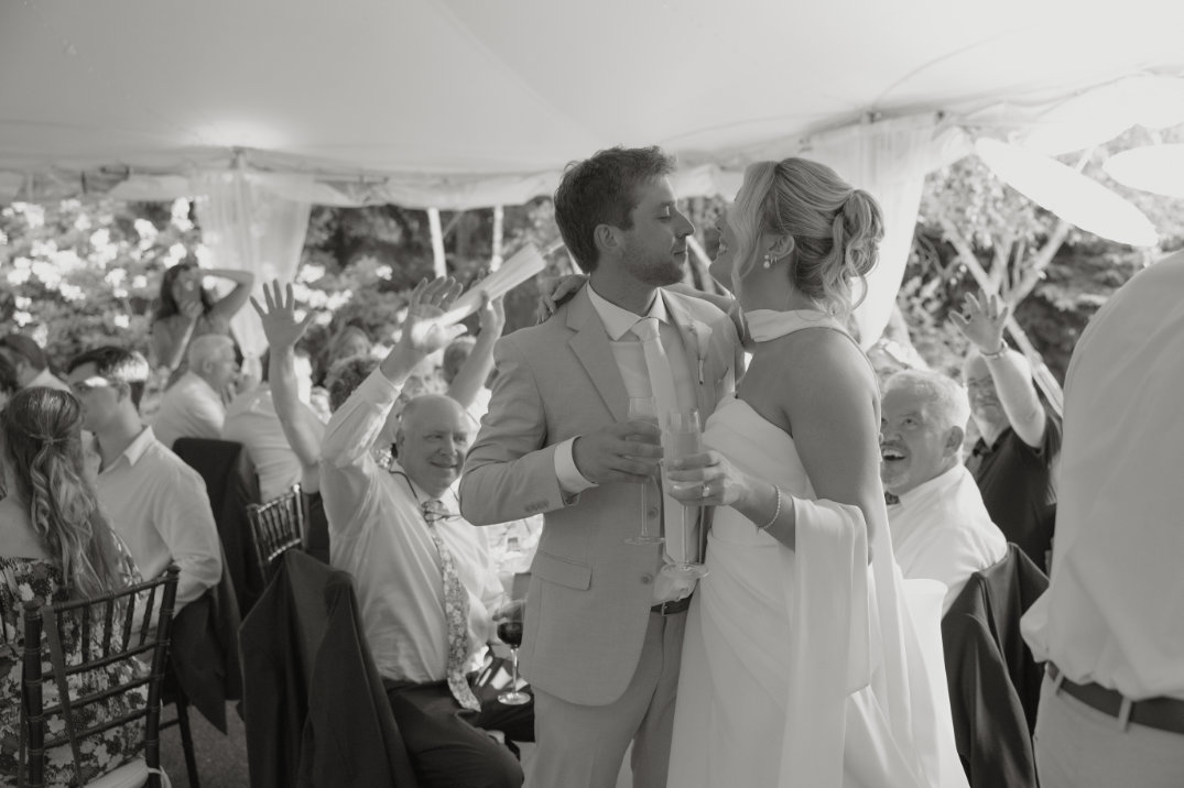 A joyful wedding scene at The Orchid House Winery shows a newlywed couple sharing a kiss. Guests in the background cheer with raised arms, celebrating the moment.