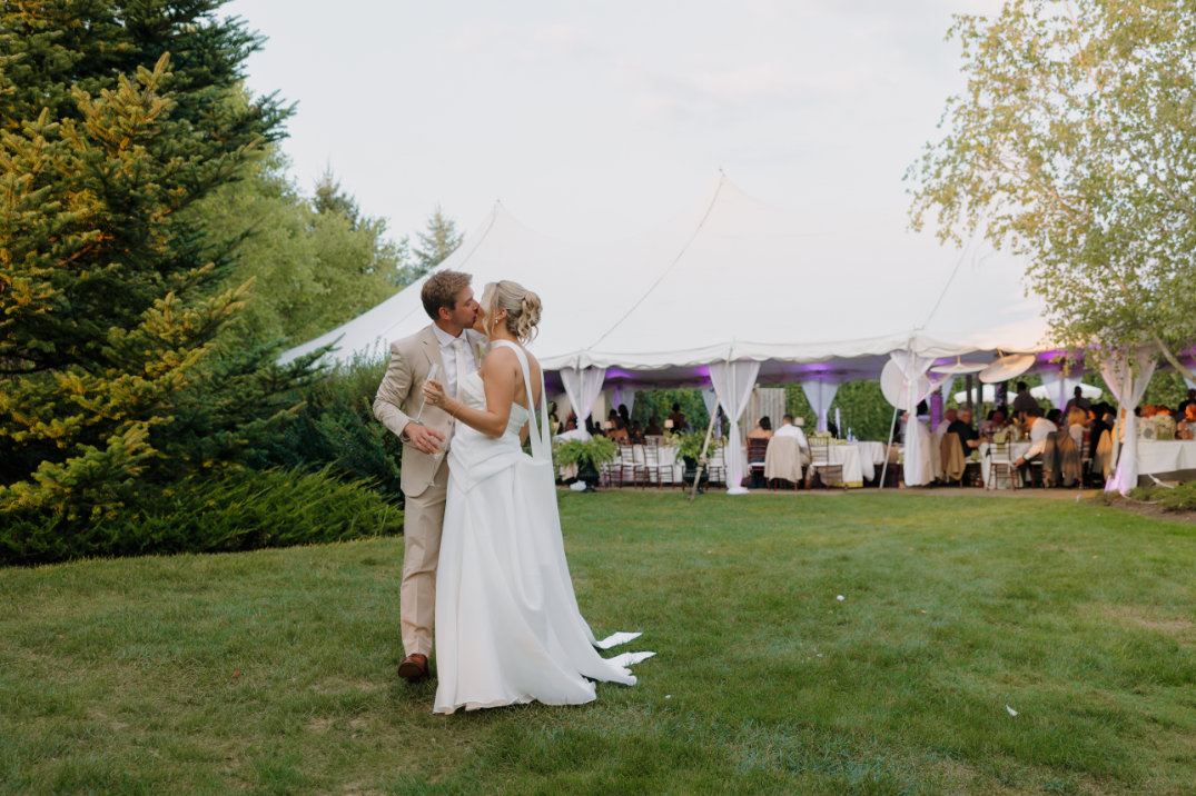 A bride in a white gown and a groom in a beige suit kiss on a grassy lawn. Behind them, a white tent with guests creates a festive wedding scene.