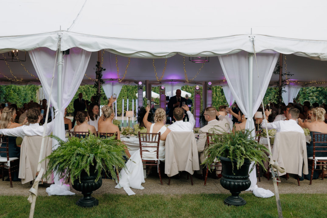 A Cleveland wedding reception under a white tent with guests seated at round tables. Elegant decor includes hanging string lights and lush greenery, creating a festive atmosphere.