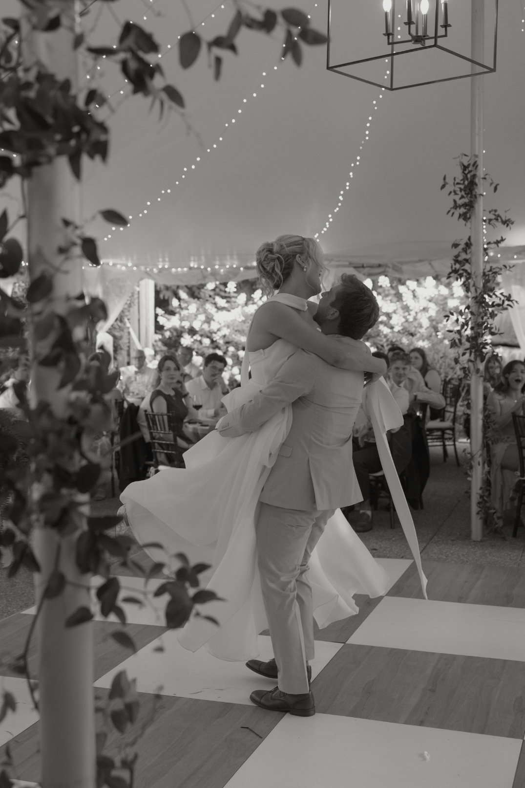A couple dances joyfully under a tent adorned with string lights. The man lifts the woman, surrounded by guests seated at decorated tables. Elegant, romantic atmosphere.