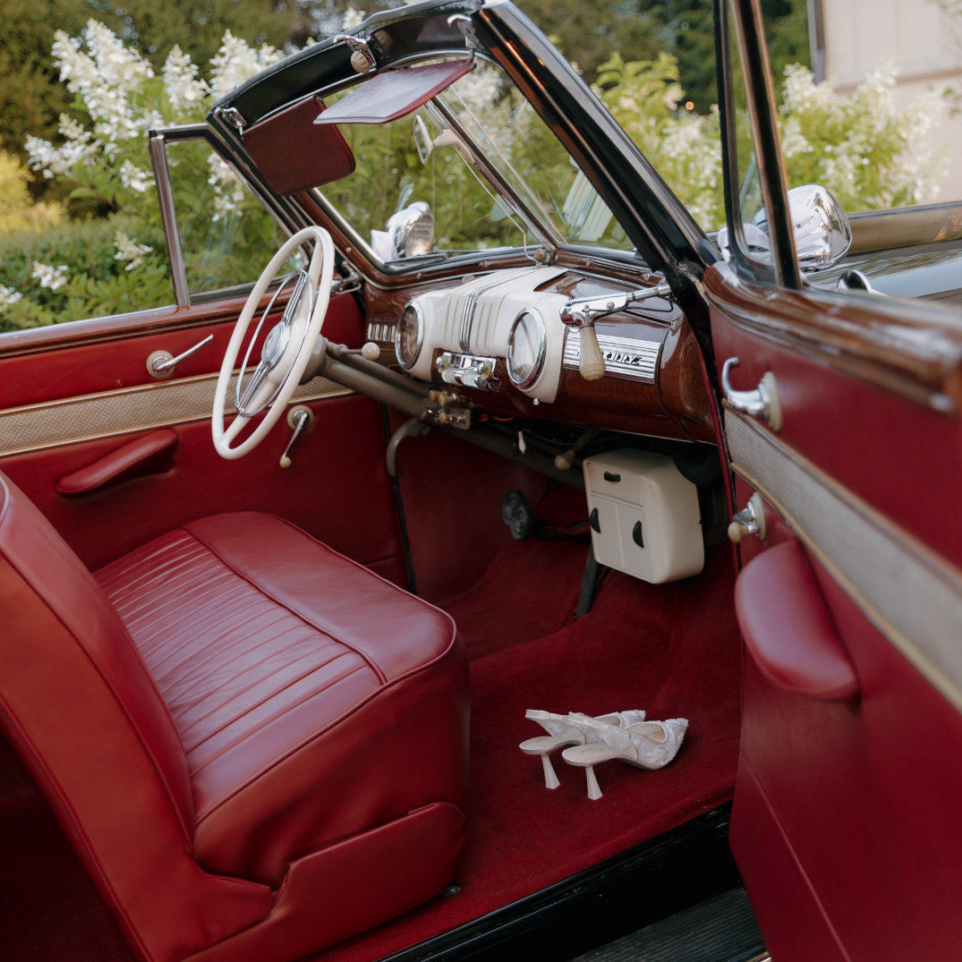 Vintage car interior with red leather seats and a classic steering wheel, set against a backdrop of green foliage. White gloves rest on the floor, evoking a nostalgic, elegant mood.