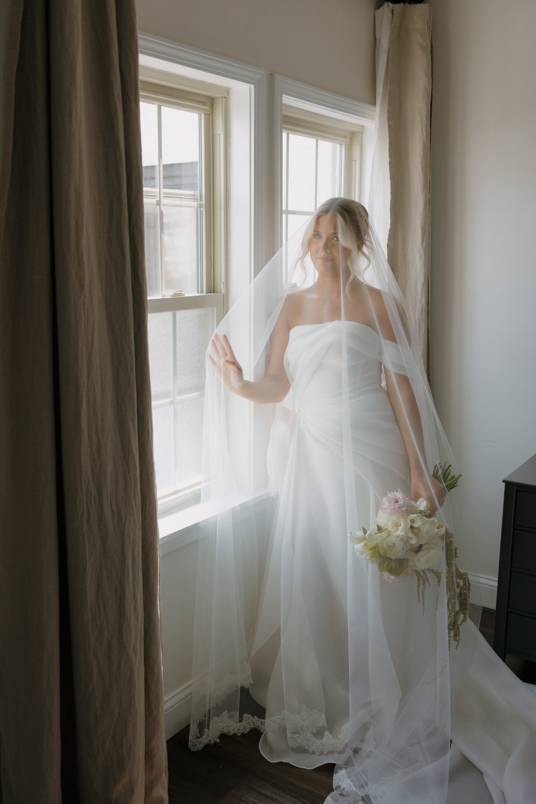 A bride in a flowing white gown and veil stands by a sunlit window holding a bouquet of flowers, creating a serene and contemplative atmosphere.