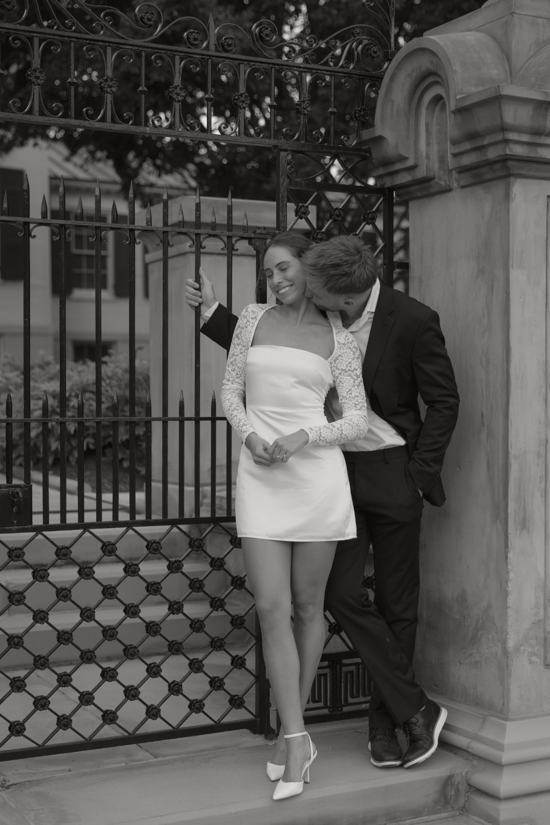 Black and white portrait of the couple standing beneath a decorative iron gate with stone architecture.