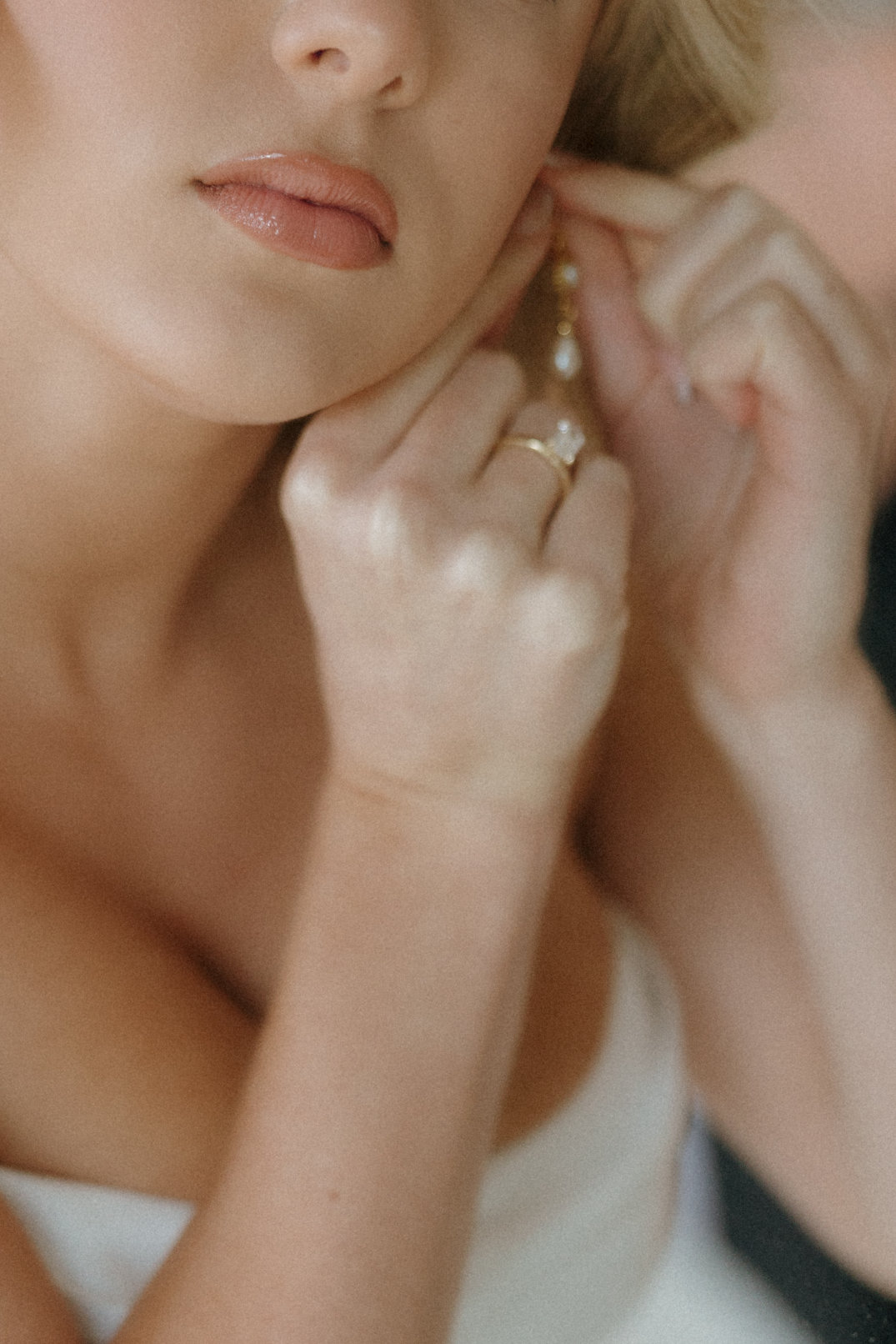 A close-up image of a woman elegantly adjusting a pearl earring, showcasing a gold ring on her finger. She wears a white strapless dress, conveying a graceful and serene mood.