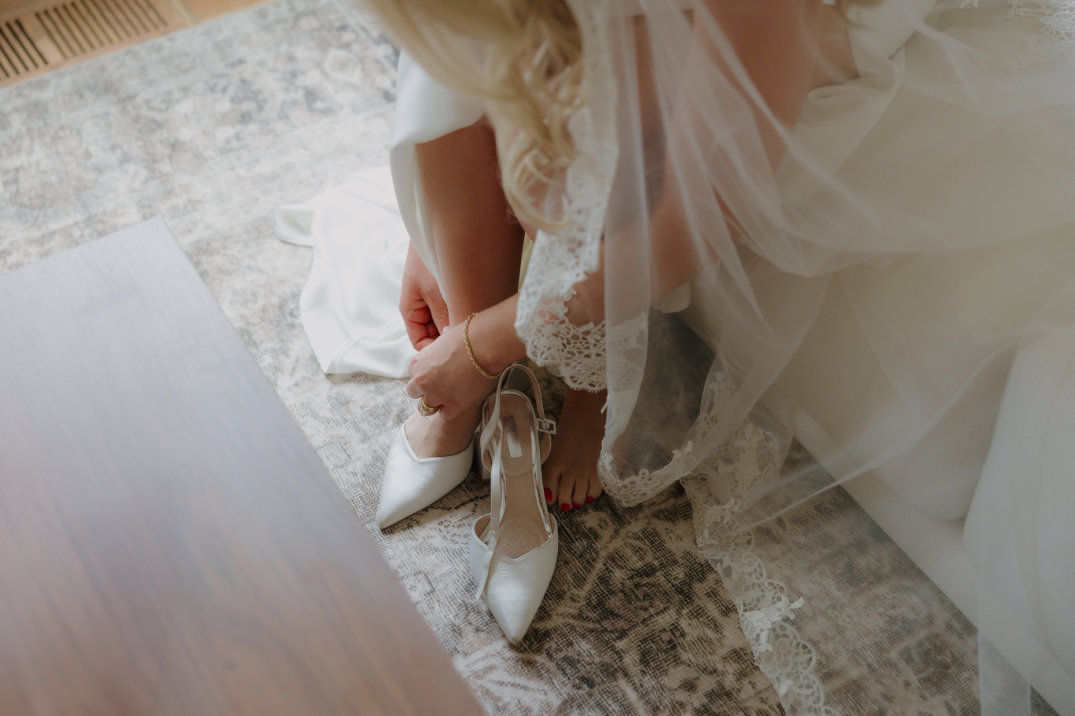Ohio bride wearing a white dress and veil kneels to adjust her white high heels on a patterned rug. The mood is intimate and serene.