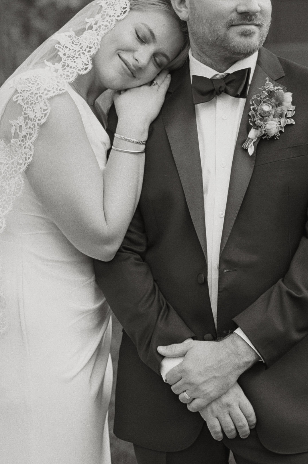 Black and white film photo of a Cincinnati bride and groom. The bride, in a lacy veil, rests her head on the groom's shoulder. The groom wears a suit with a floral boutonniere. Peaceful and intimate.