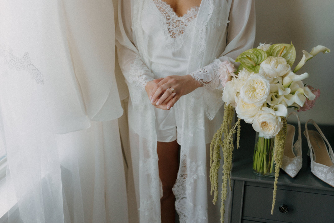 Ohio Bride in a lacy white robe stands by a window, holding hands. Beside her, a bouquet of white flowers and wedding shoes on a gray dresser.