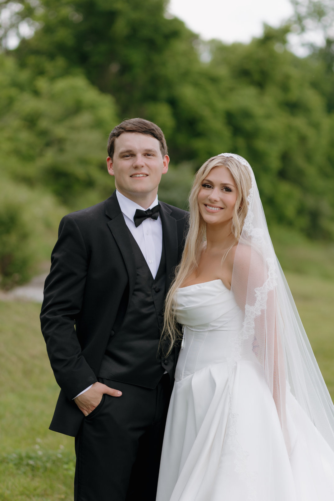 Portrait of the bride and groom standing close together, smiling softly toward the camera in a relaxed outdoor setting.