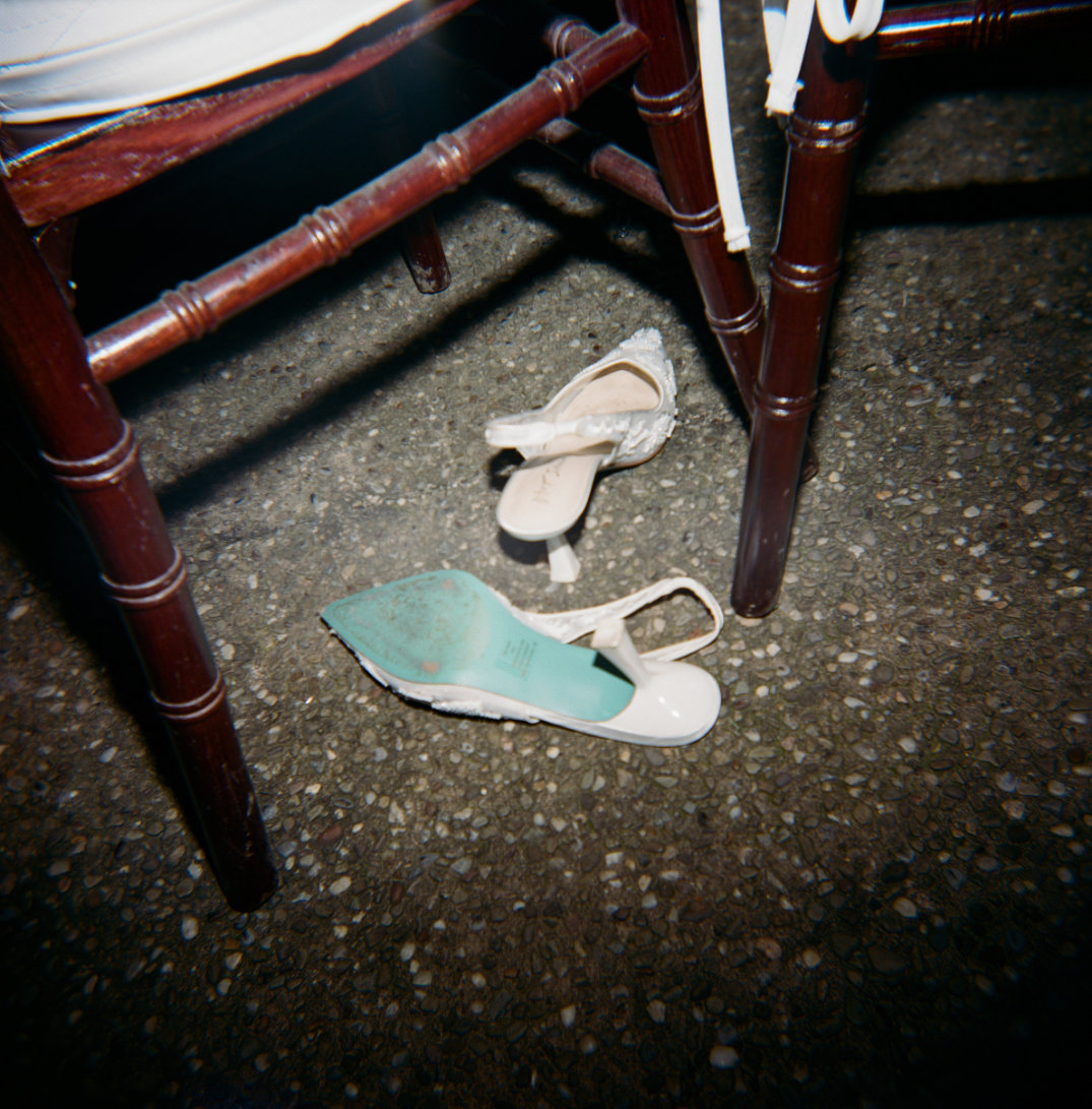 White high-heeled shoes, slightly worn, rest under a dark wooden chair on a stone floor, suggesting a relaxed or informal setting.