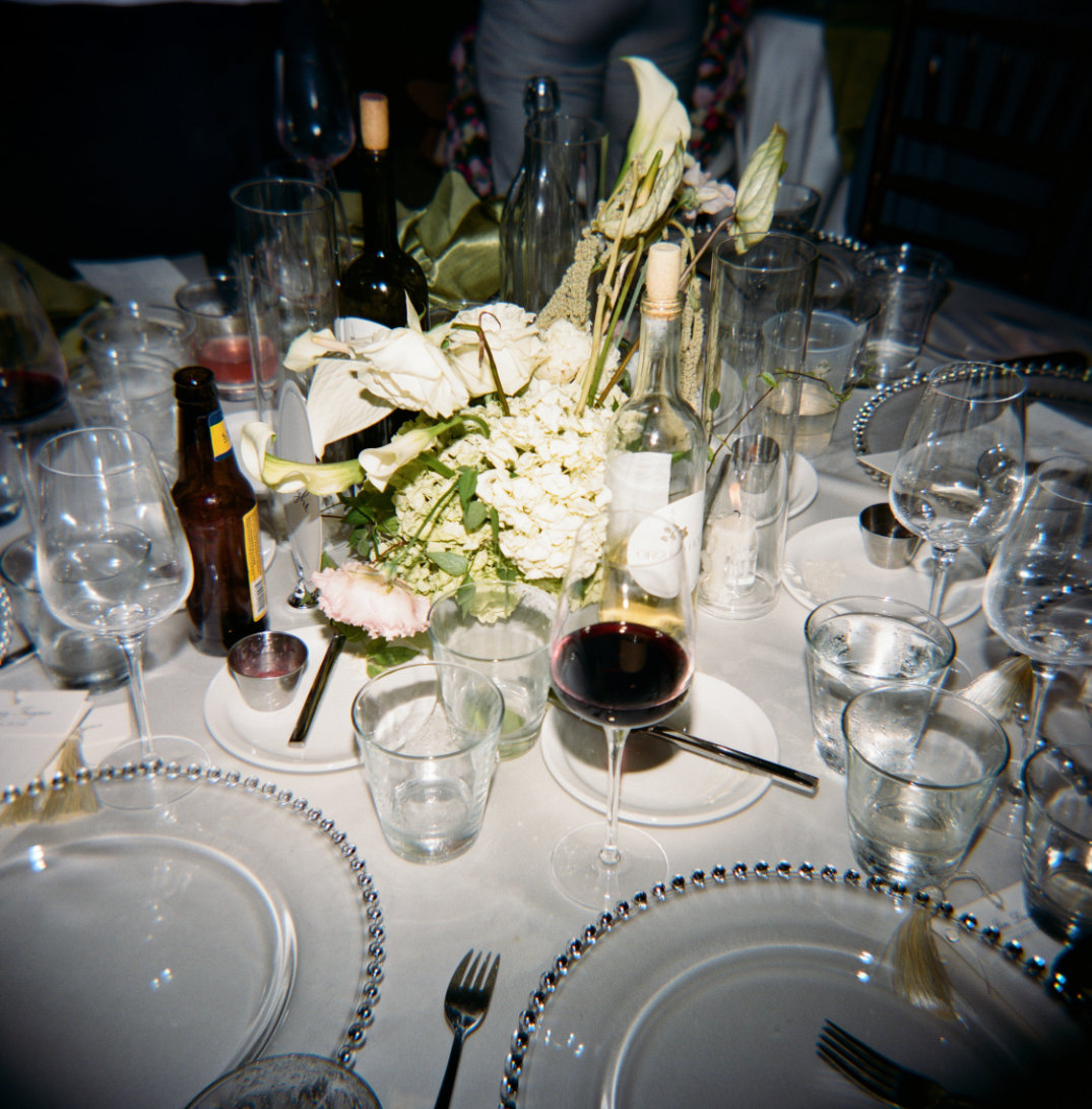 Elegant dinner table with white floral centerpiece, surrounded by empty wine glasses, bottled wine, and clear plates with beaded rims taken with a film camera in Ohio.