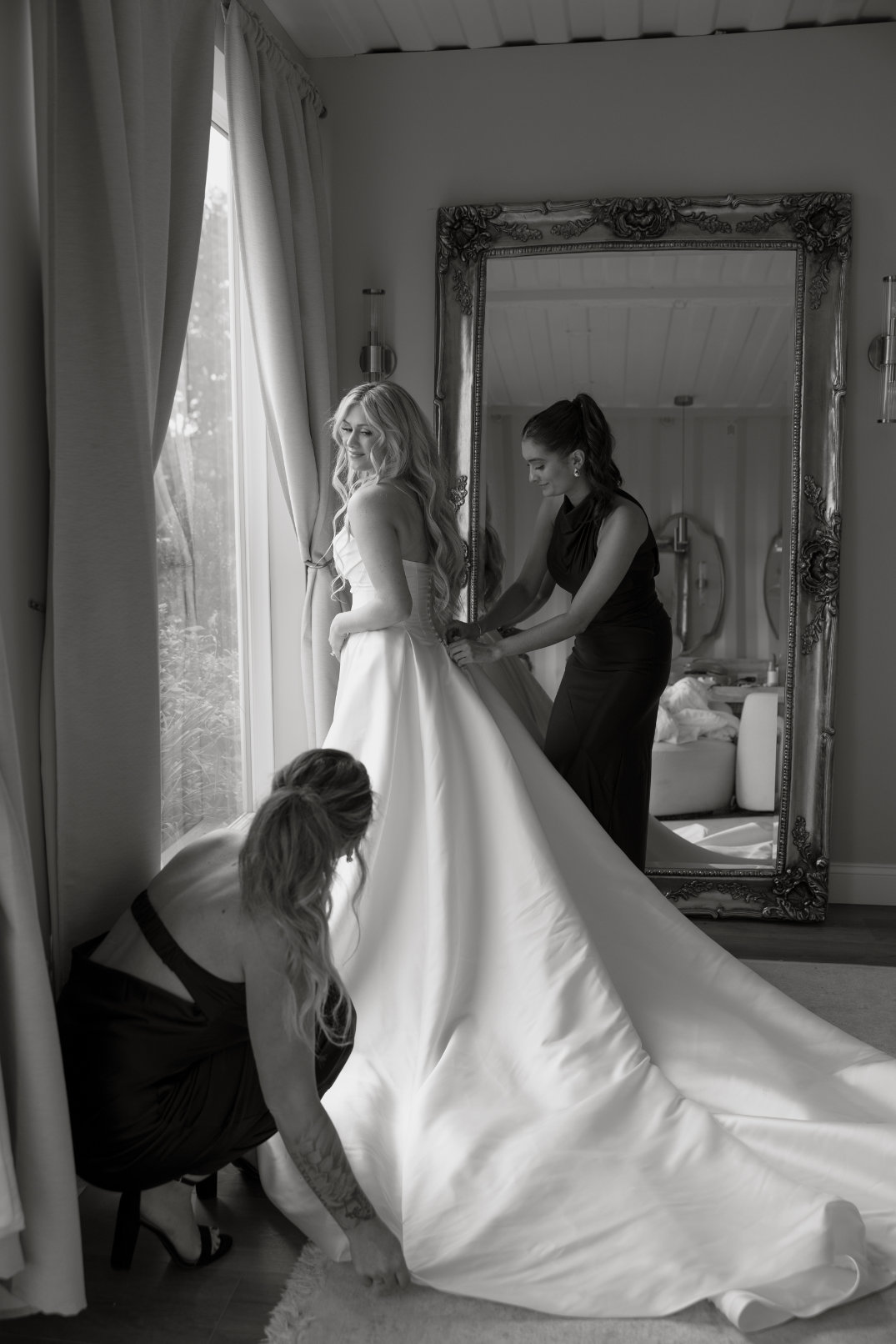 A black-and-white film image shows an Ohio bride smiling gently, assisted by two women adjusting her elegant gown. A large ornate mirror reflects the scene.