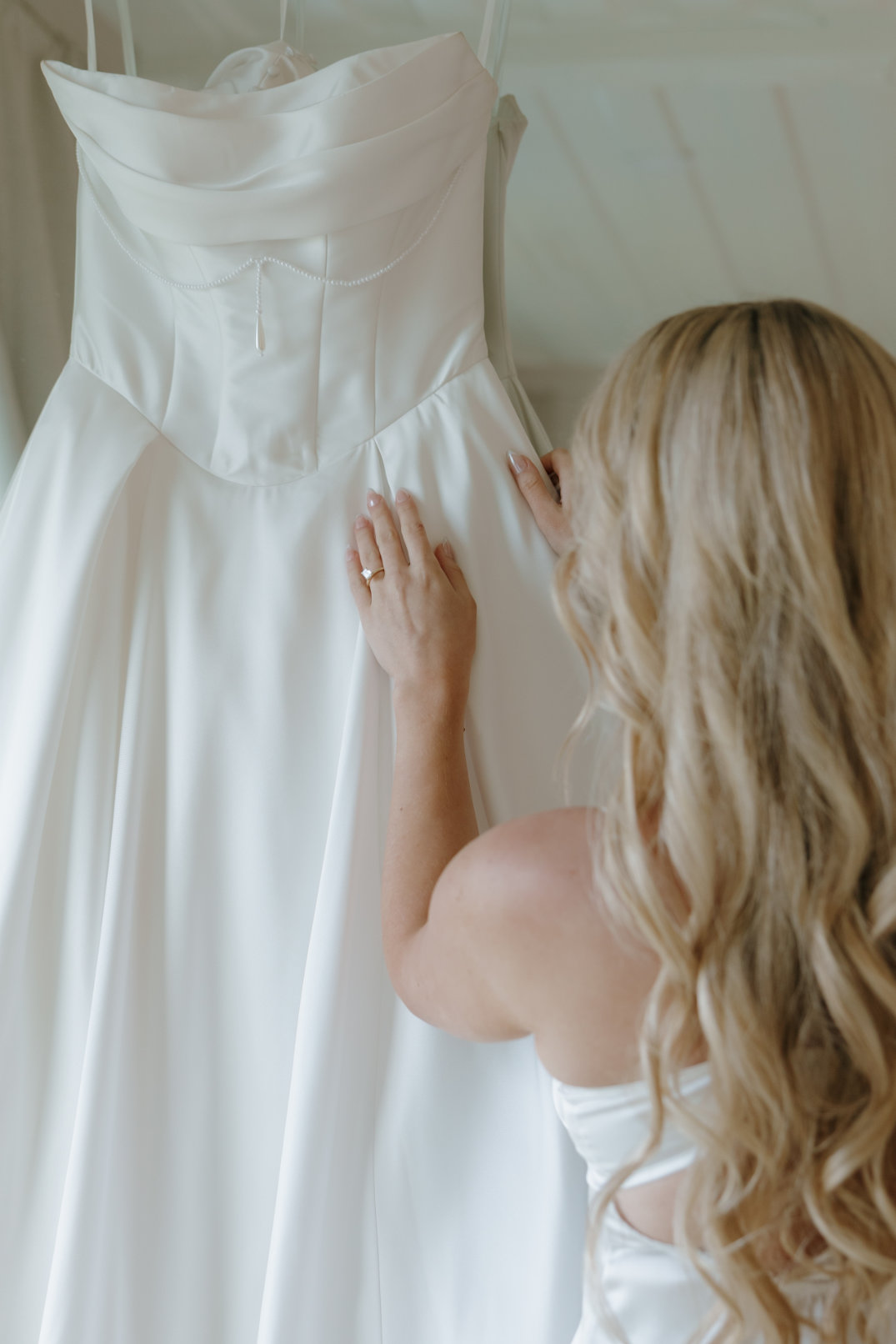 A woman with long blonde hair touches a hanging white wedding dress in a softly lit room. The scene conveys anticipation and elegance.