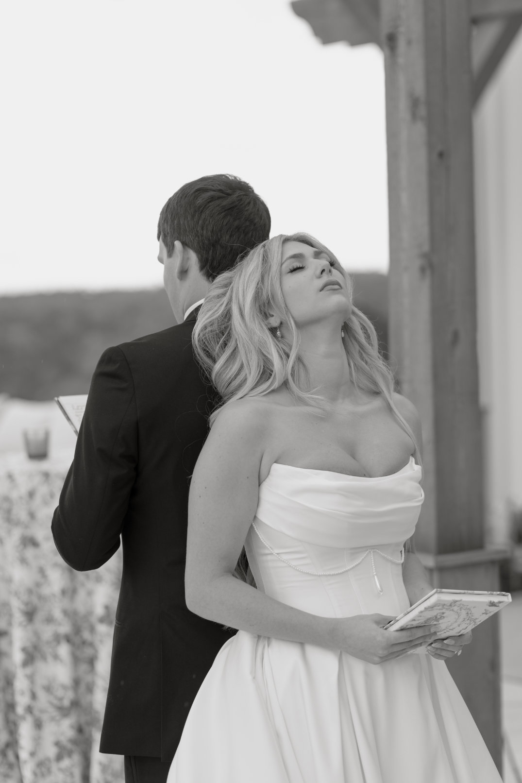 A bride and groom stand back-to-back, holding books, creating an emotional moment. The bride looks thoughtful, wearing a strapless gown.
