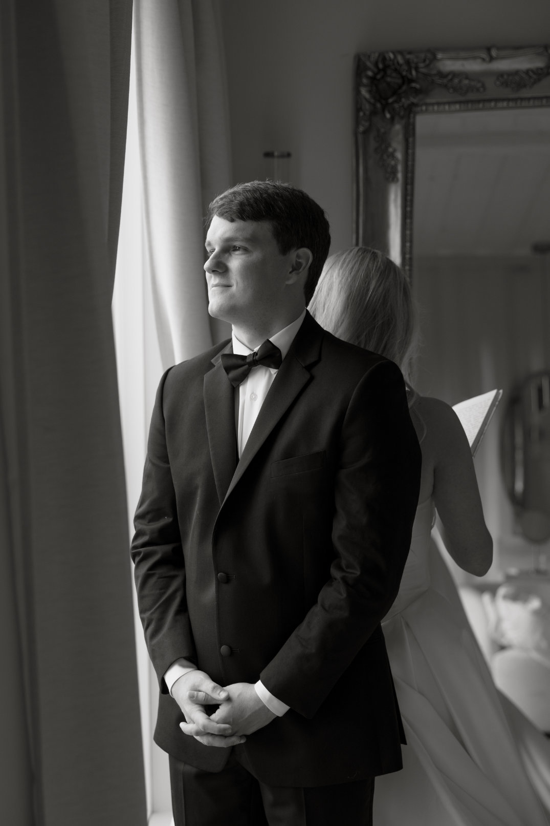 A groom in a black tuxedo stands in a room by a window, hands clasped, looking serene. A bride in a white dress stands behind him, reading a letter.