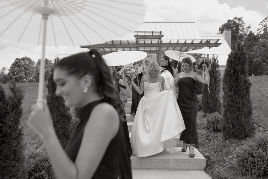 Black and white image of a joyful bridal party descending stone steps in Hocking Hills. The bride in a gown holds an umbrella, surrounded by smiling bridesmaids. Elegant, celebratory ambiance.