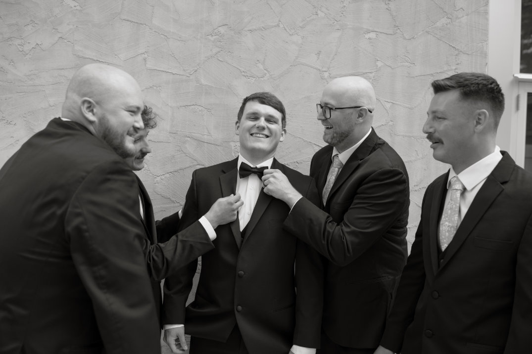 Five men in suits share a joyful moment, adjusting a central man's bow tie. The mood is playful and celebratory, with a textured wall behind them.