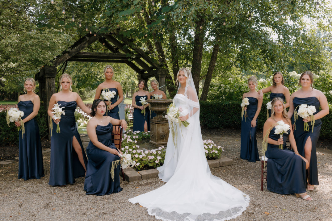 Bride in a white gown stands with bridesmaids in navy dresses holding white bouquets. The group poses outdoors amid greenery, exuding elegance and joy.