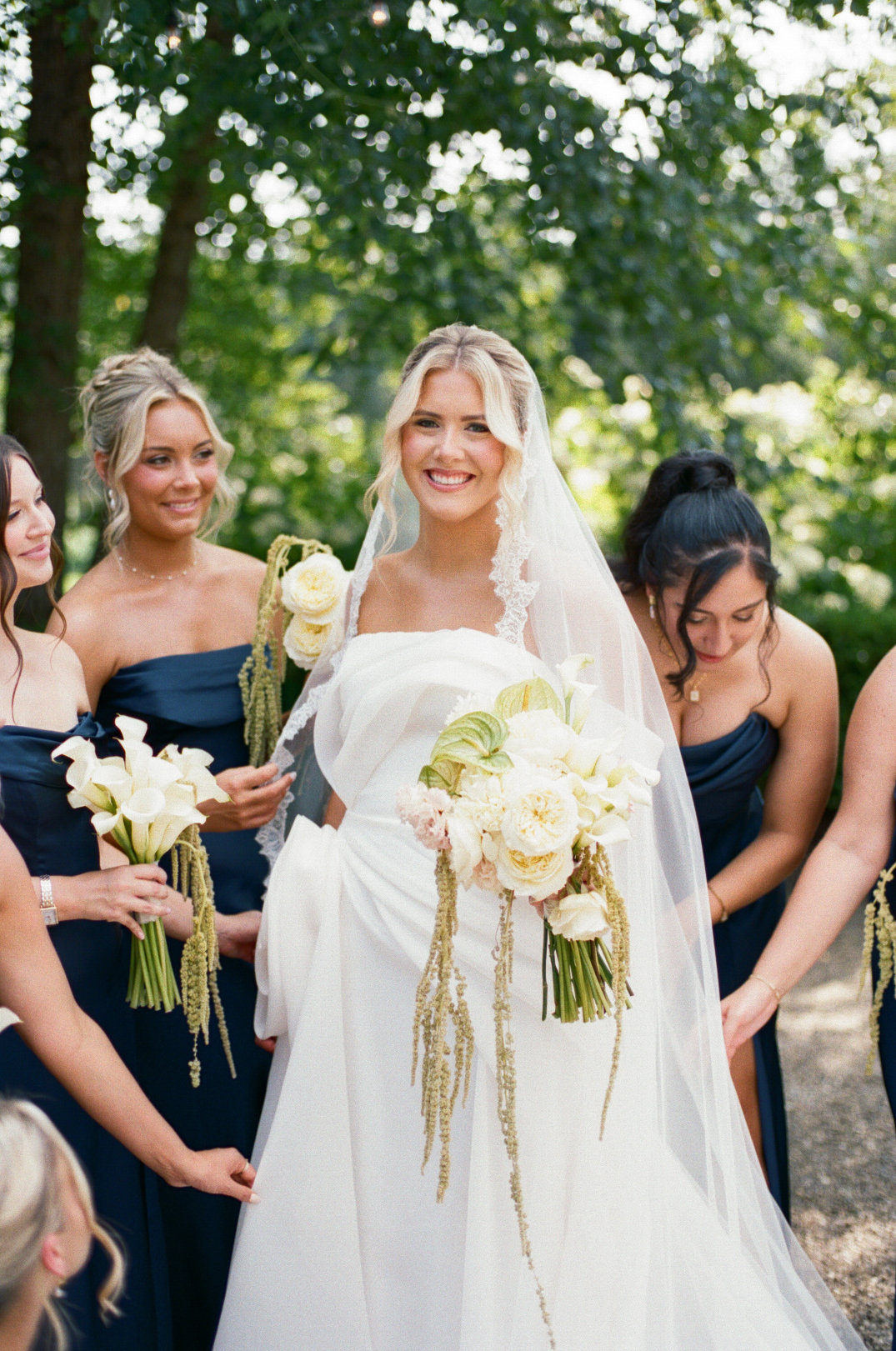 A joyful bride in a white gown and veil holds a bouquet of white flowers, surrounded by bridesmaids in navy dresses, smiling in a lush, green garden at the Orchid House Winery, Cleveland, Ohio