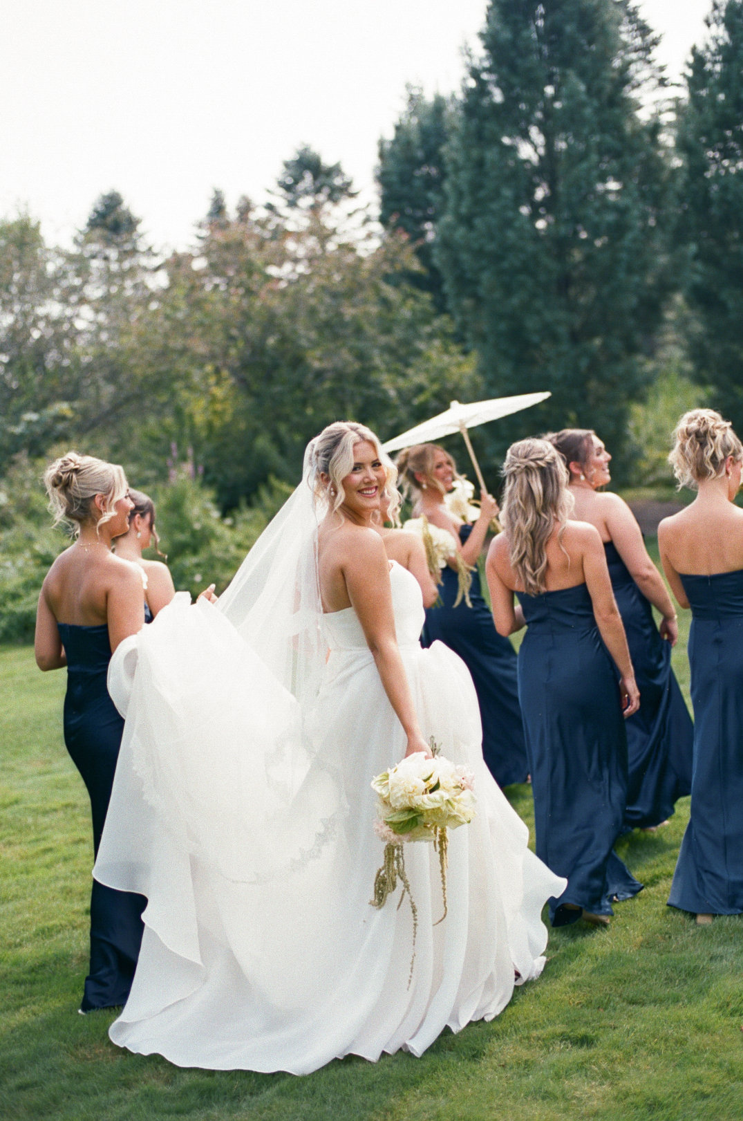 Cleveland, Ohio Bride in a white dress smiles joyfully, surrounded by bridesmaids in navy dresses holding a white parasol, with a lush garden background.